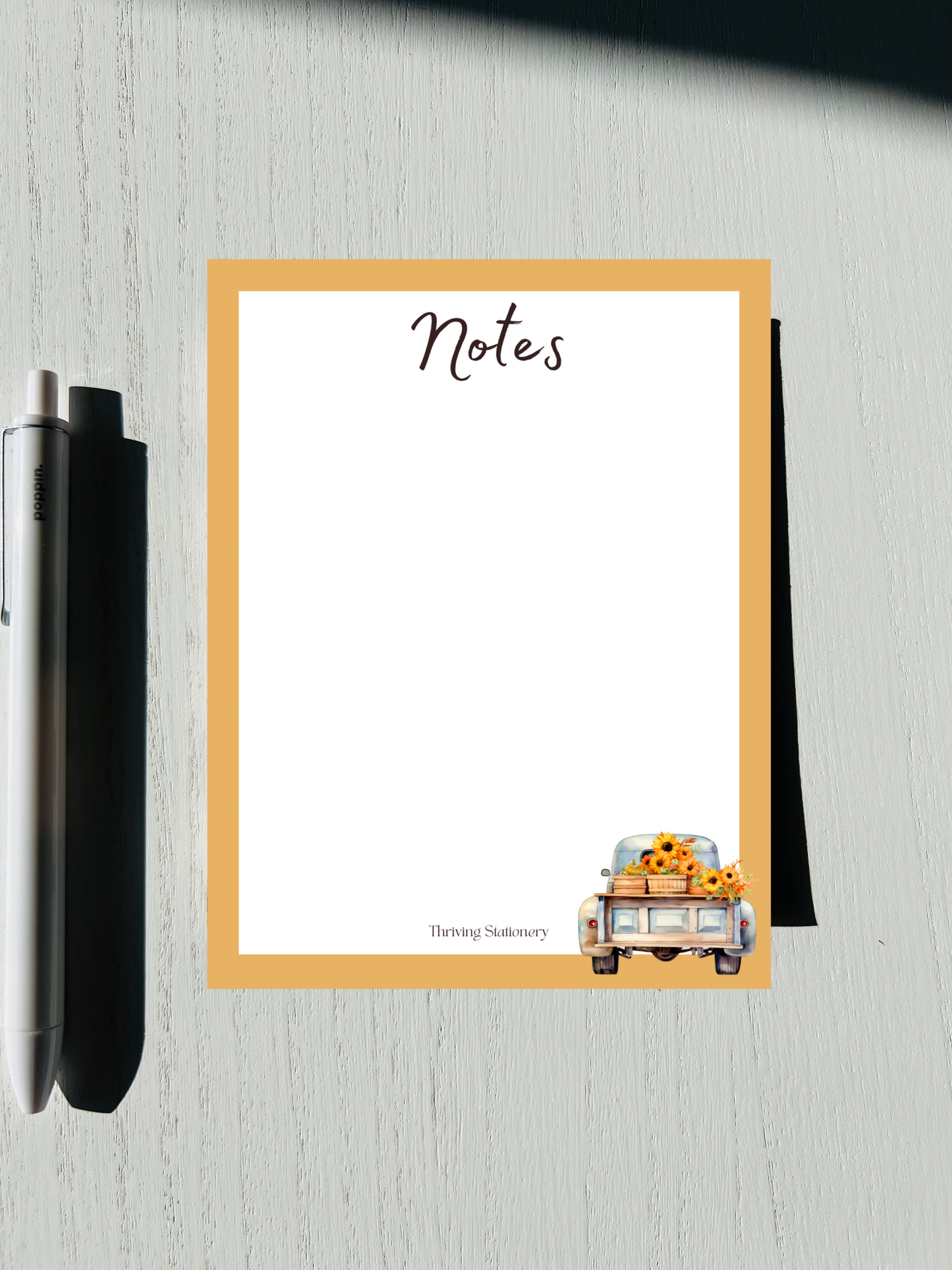 A notepad , featuring a design with autumn-themed graphics such as leaves and a pumpkin, alongside a pen and placed on a wooden surface.