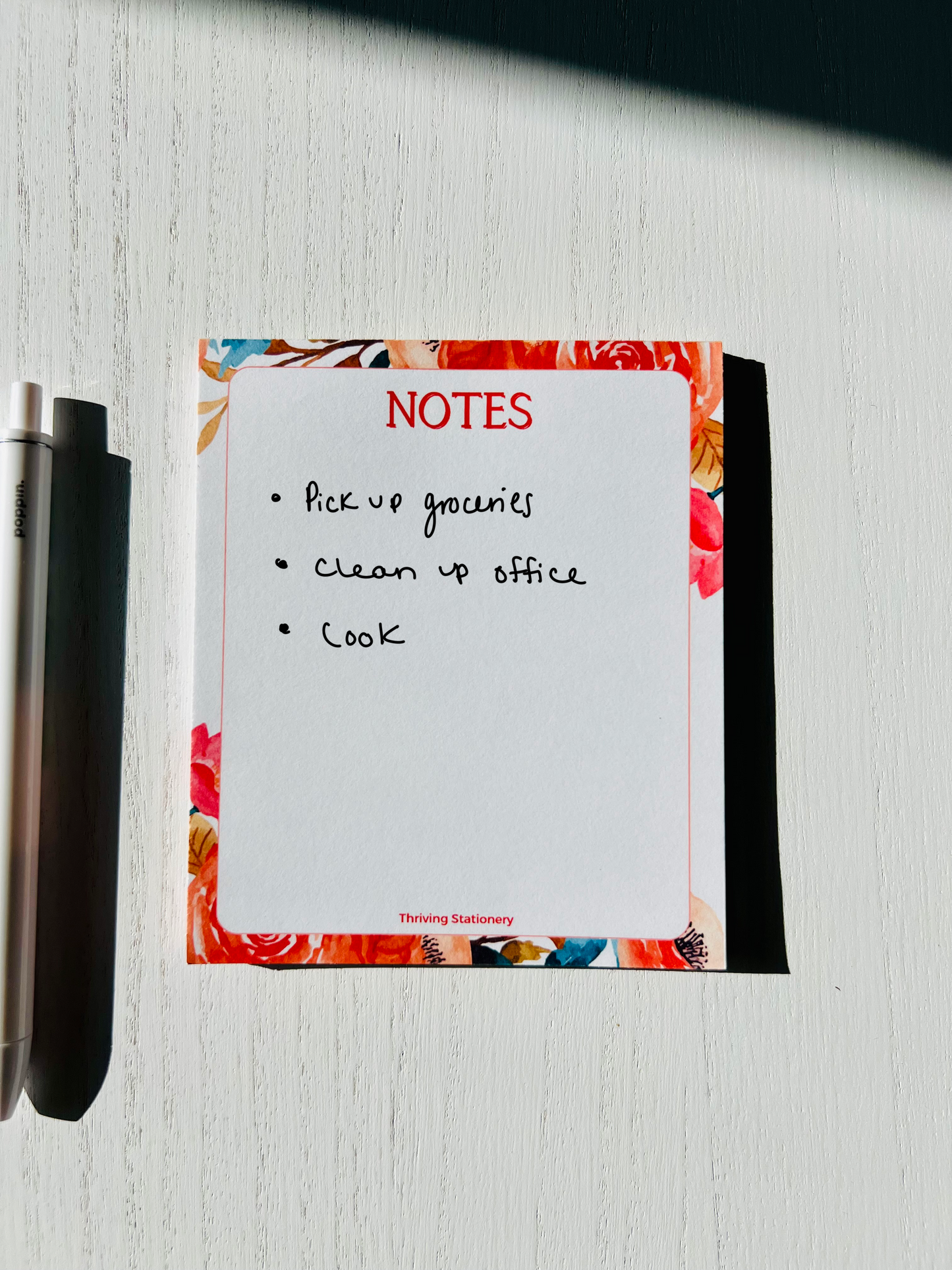 A mini notepad with a white, red, and natural color scheme, featuring a floral design on the cover, with a pen beside it.