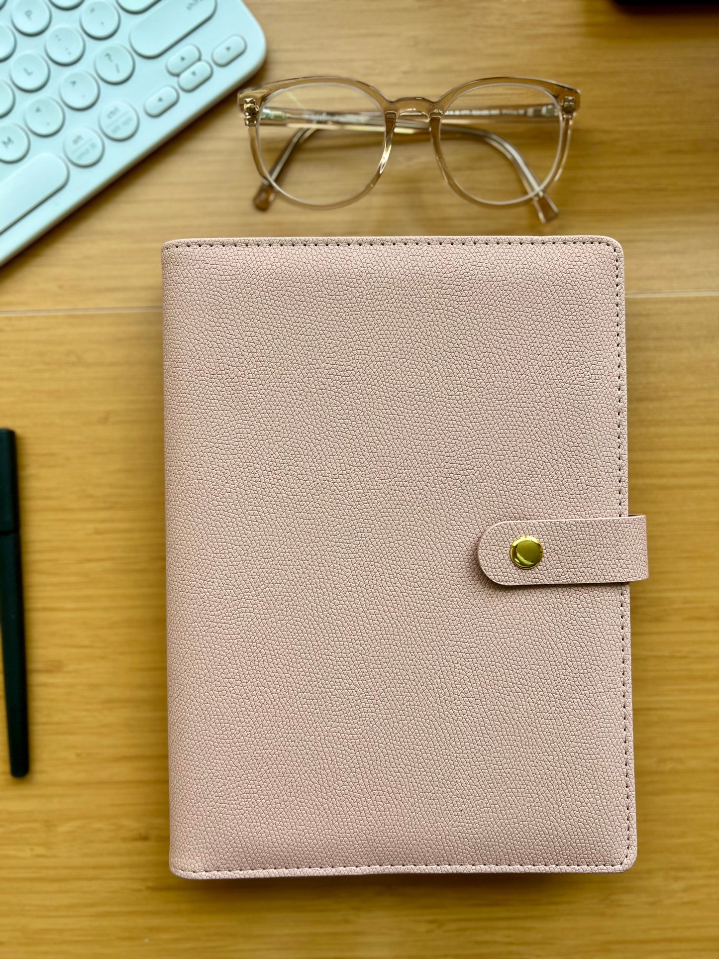 Beige planner with a gold buckle on a wooden desk with glasses and a keyboard.
