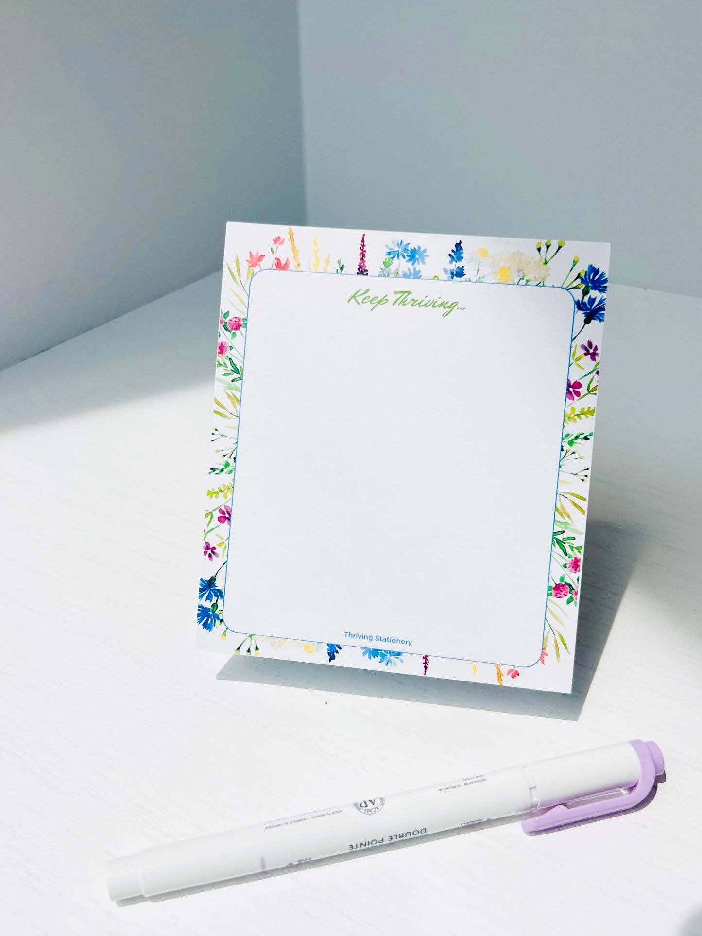 Floral-bordered notepad with “Keep Thriving…” at the top and vibrant watercolor flowers around a blank center. Logo at bottom reads “Thriving Stationery.”