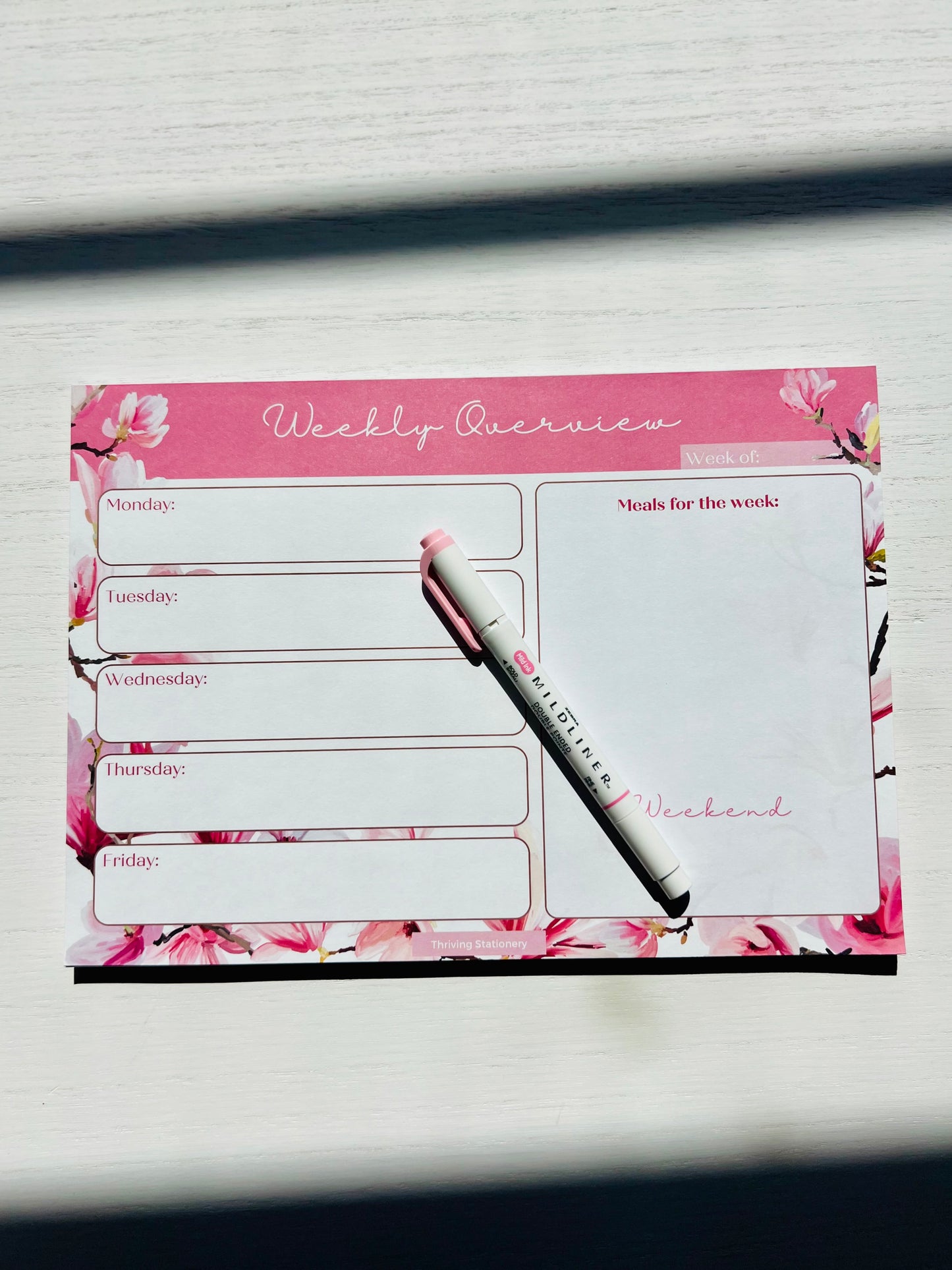 A pink weekly planner desk pad with floral borders and cherry blossom designs, featuring a white pen and marked with 'Weekly Review' in a flower-like font on the cover.