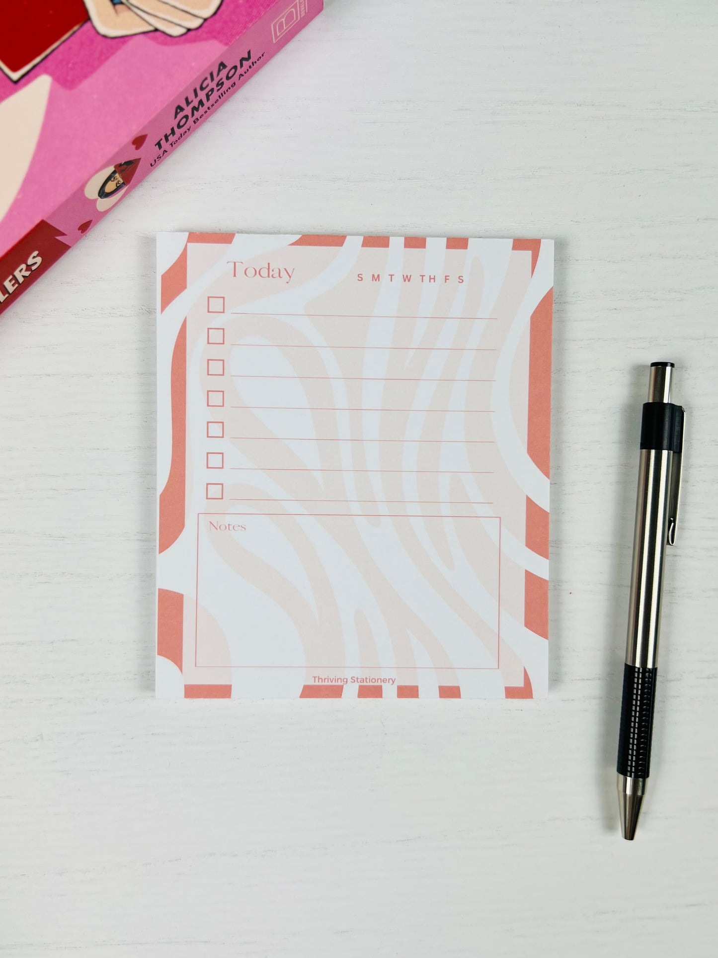 Coral Flow Daily Planner Notepad with coral and cream wavy border, checklist, and notes section with book and pen for scale