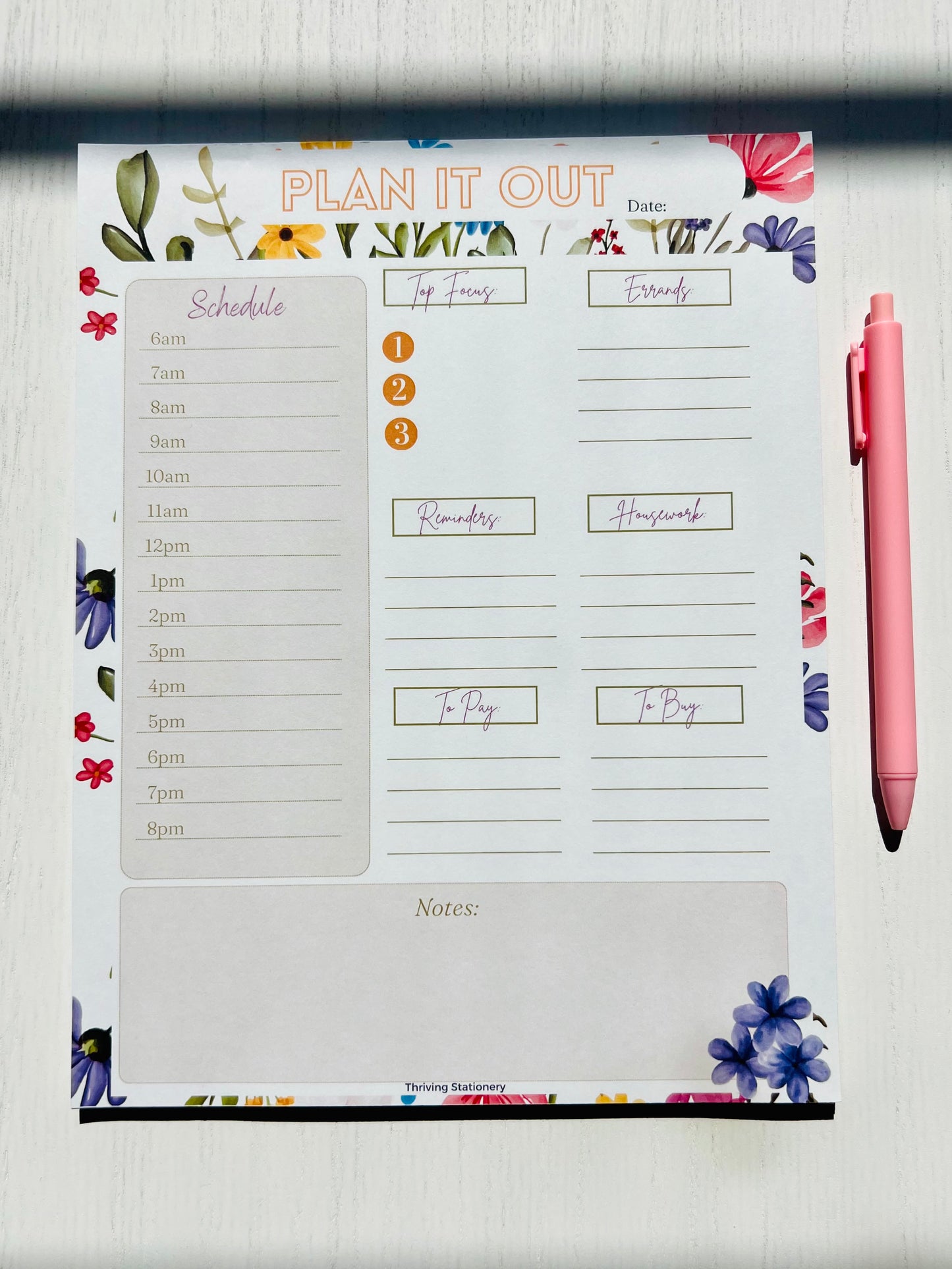 A white daily planner notepad with a floral design on the pages, accompanied by a pink pen.