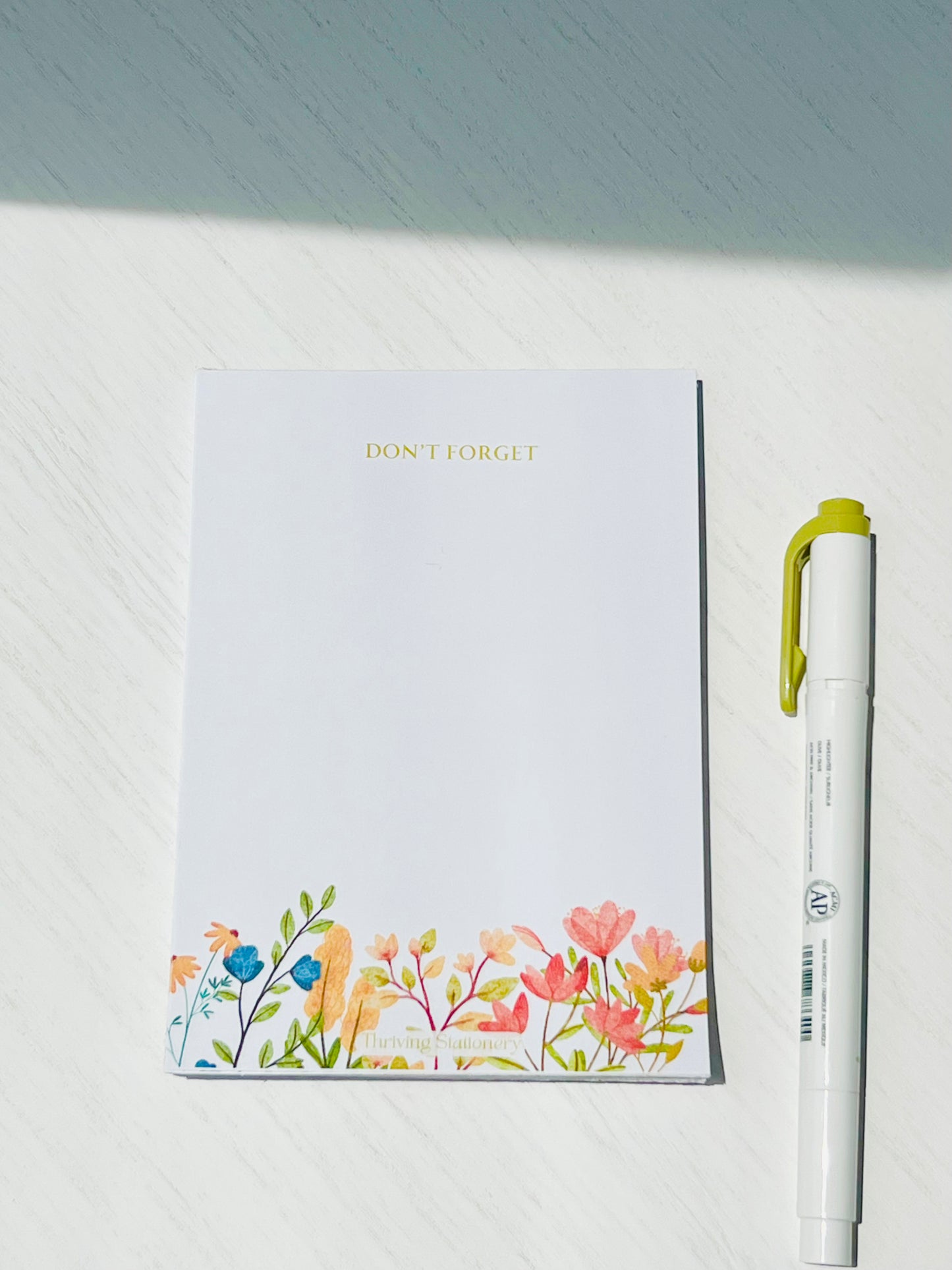 A white notepad with a vibrant late summer blooms design on the cover, accompanied by a green pen and a yellow highlighter.