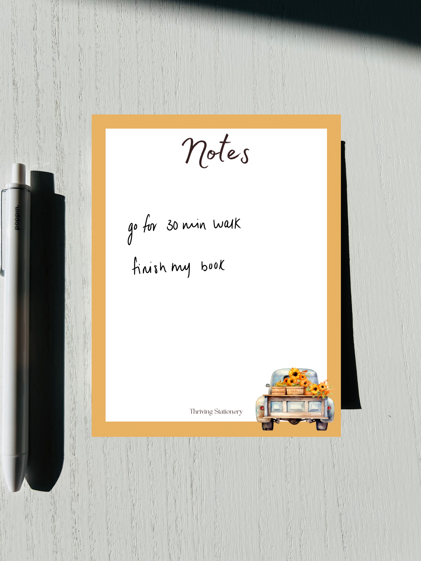 A notepad written, featuring a design with autumn-themed graphics such as leaves and a pumpkin, alongside a pen and placed on a wooden surface.