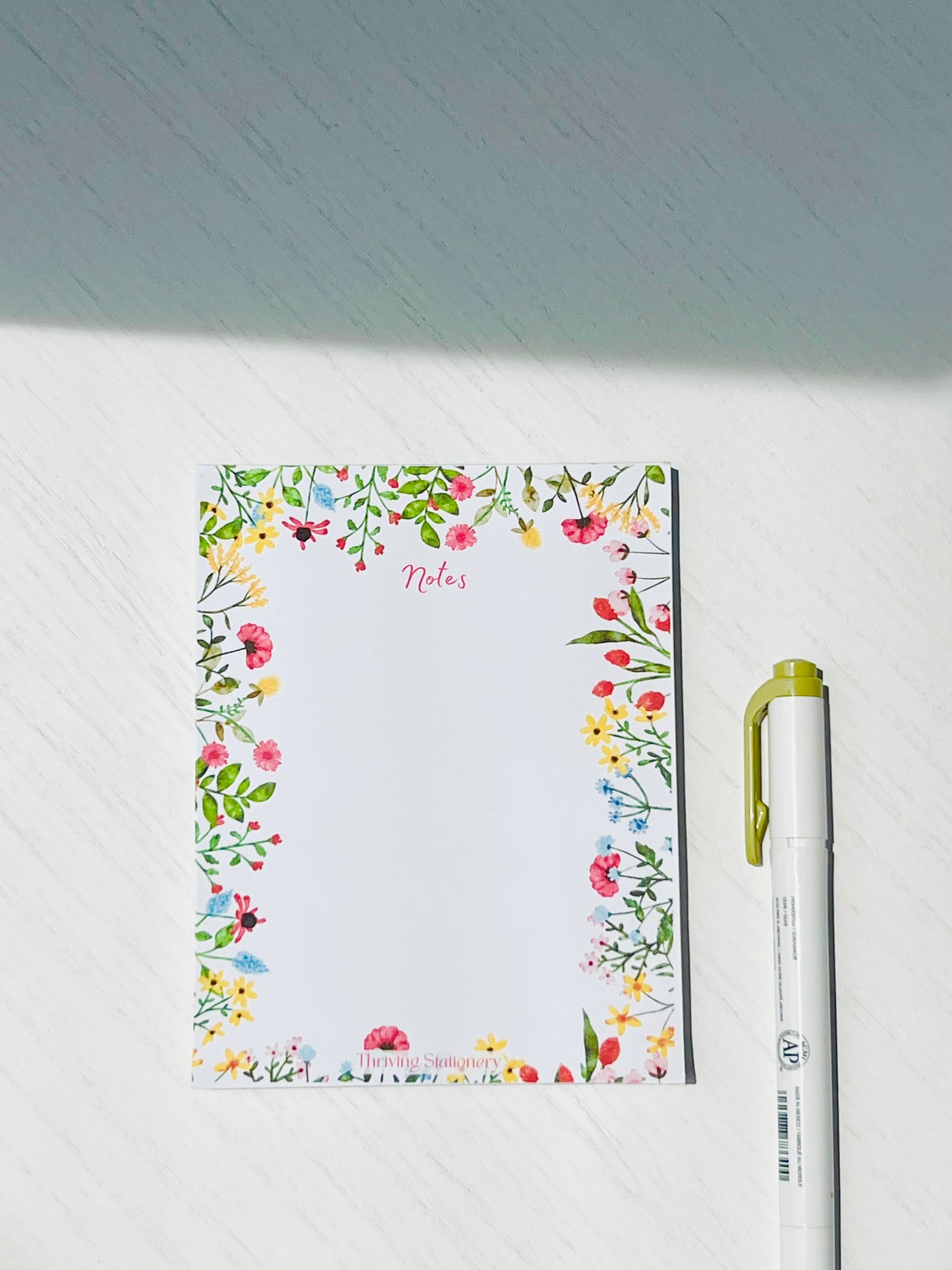 A floral-themed blank notepad with a pen beside it, featuring a garden design with various flowers and a space for writing.