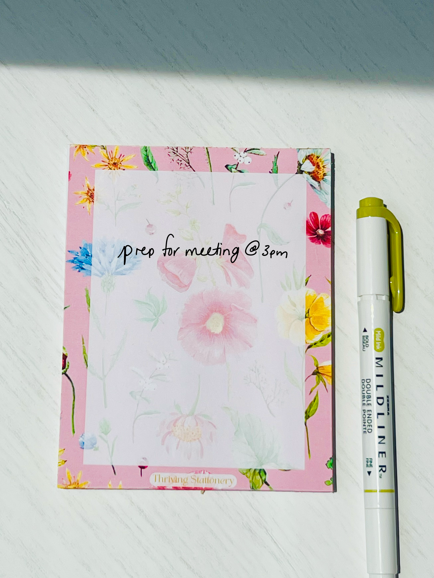Floral notepad with soft watercolor wildflowers on a blush pink background. “Thriving Stationery” logo at the bottom center.