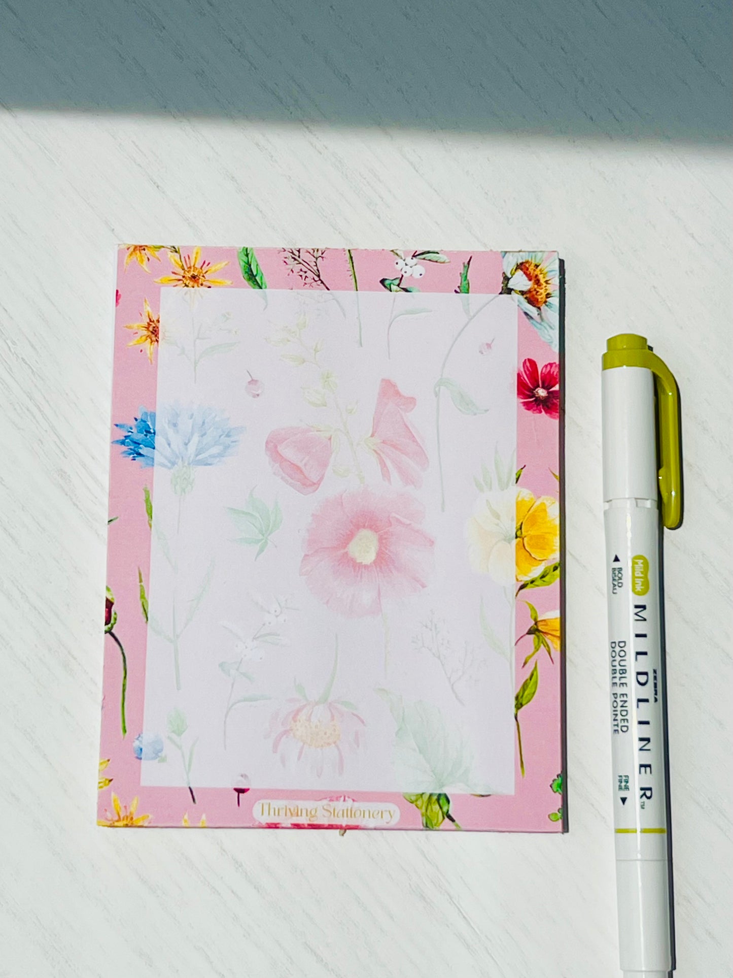 Floral notepad with soft watercolor wildflowers on a blush pink background. “Thriving Stationery” logo at the bottom center.
