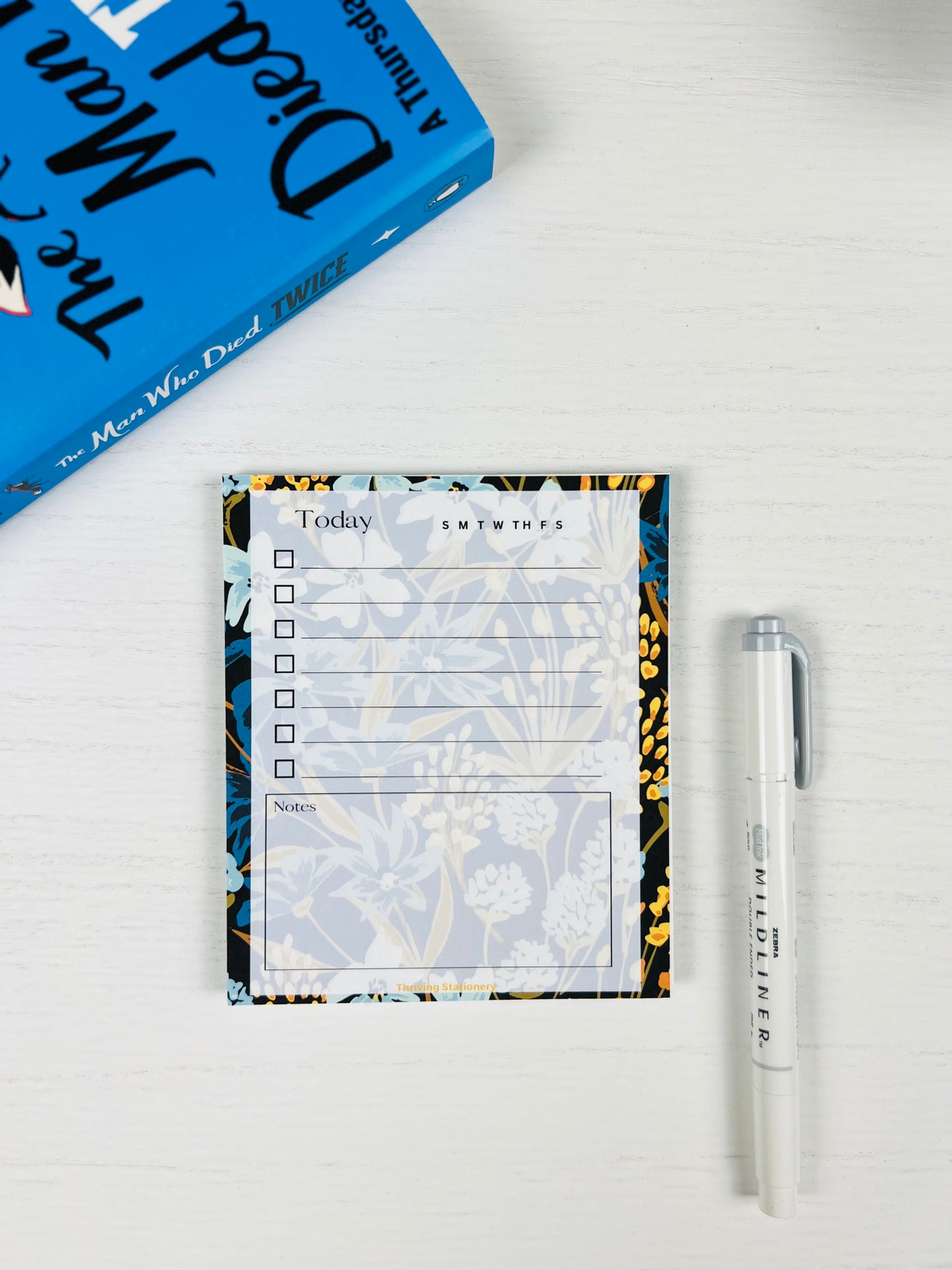 Navy Meadow Daily Planner Notepad with navy, blue, and gold floral border and daily checklist layout with a book and pen for scale