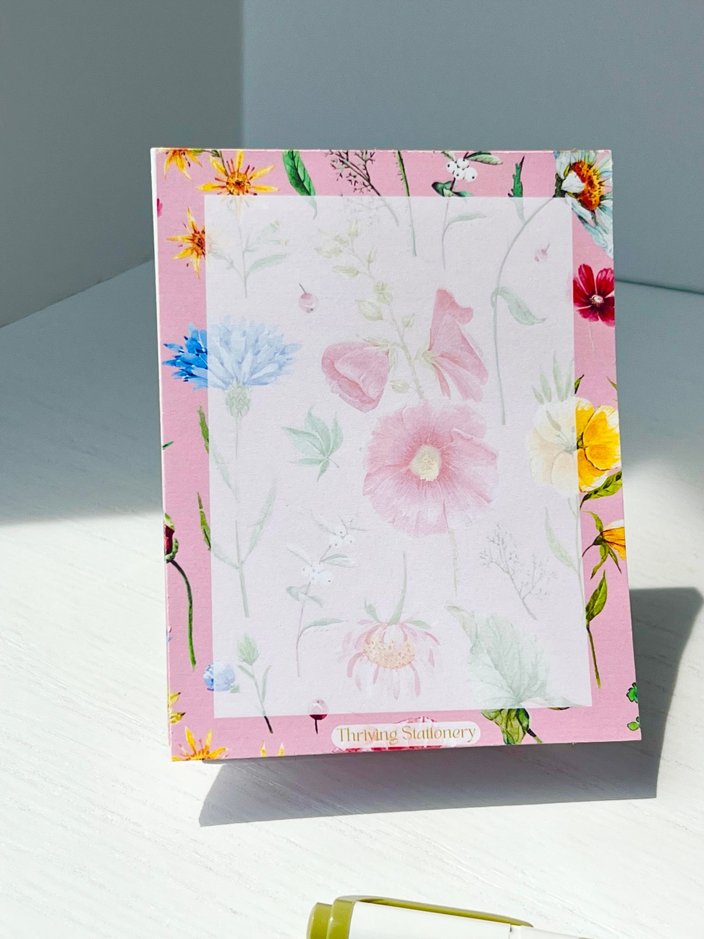 Floral notepad with soft watercolor wildflowers on a blush pink background. “Thriving Stationery” logo at the bottom center.