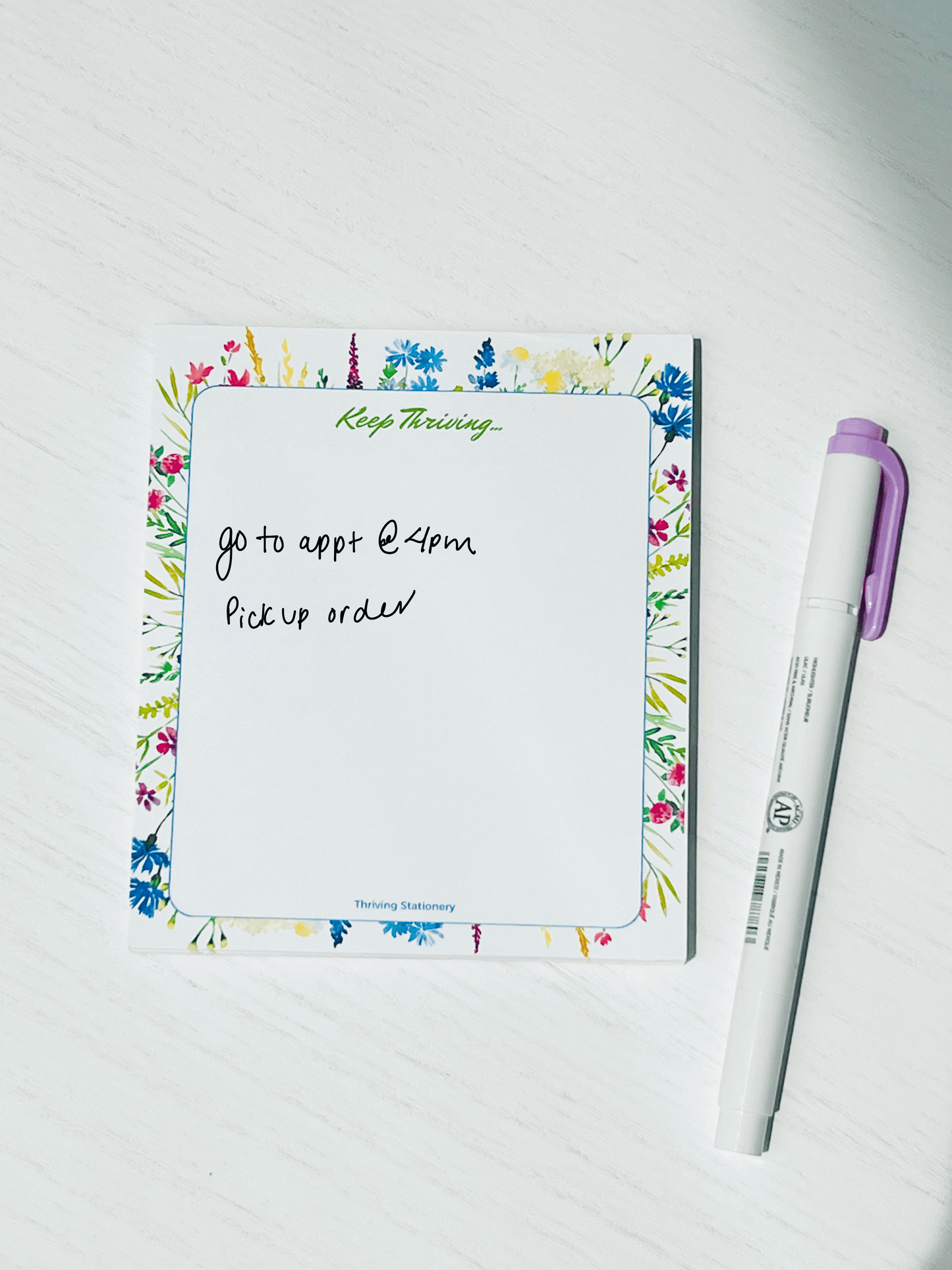 Floral-bordered notepad with “Keep Thriving…” at the top and vibrant watercolor flowers around a blank center. Logo at bottom reads “Thriving Stationery.”
