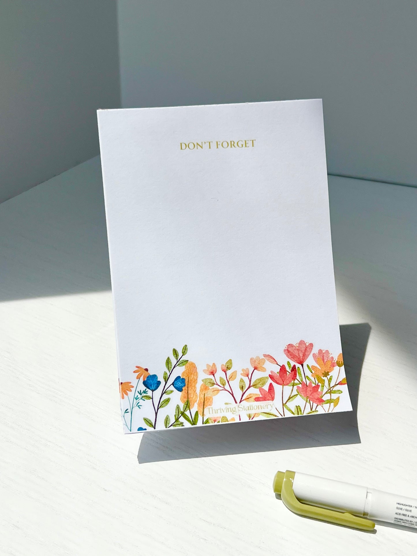 Minimal white notepad with soft watercolor floral illustrations along the bottom and “DON’T FORGET” in gold text at the top. Branding reads “Thriving Stationery.