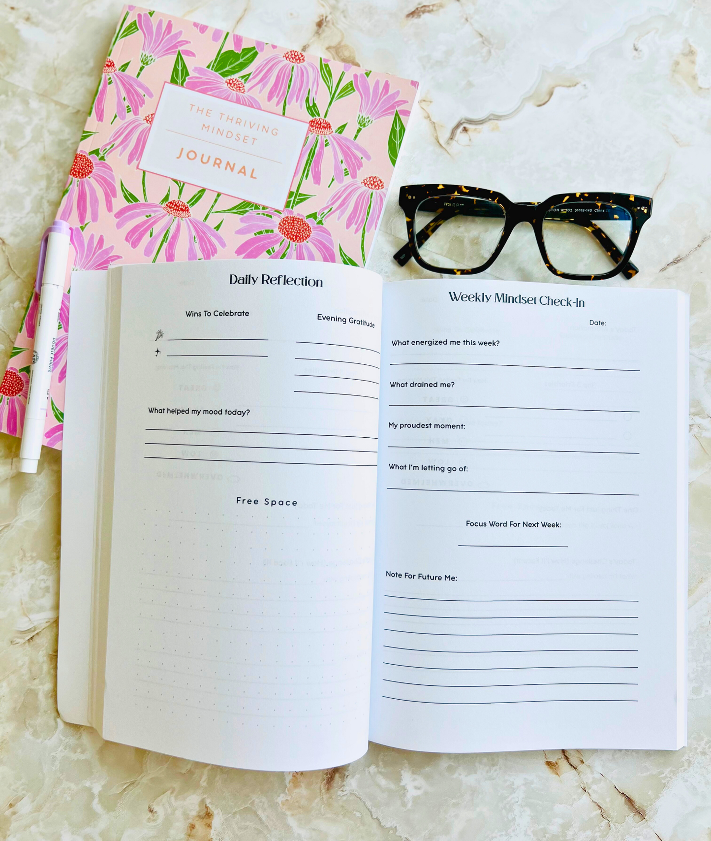 The Thriving Mindset Journal with pink floral cover and 90-day guided journaling layout with glasses and a pen for scale