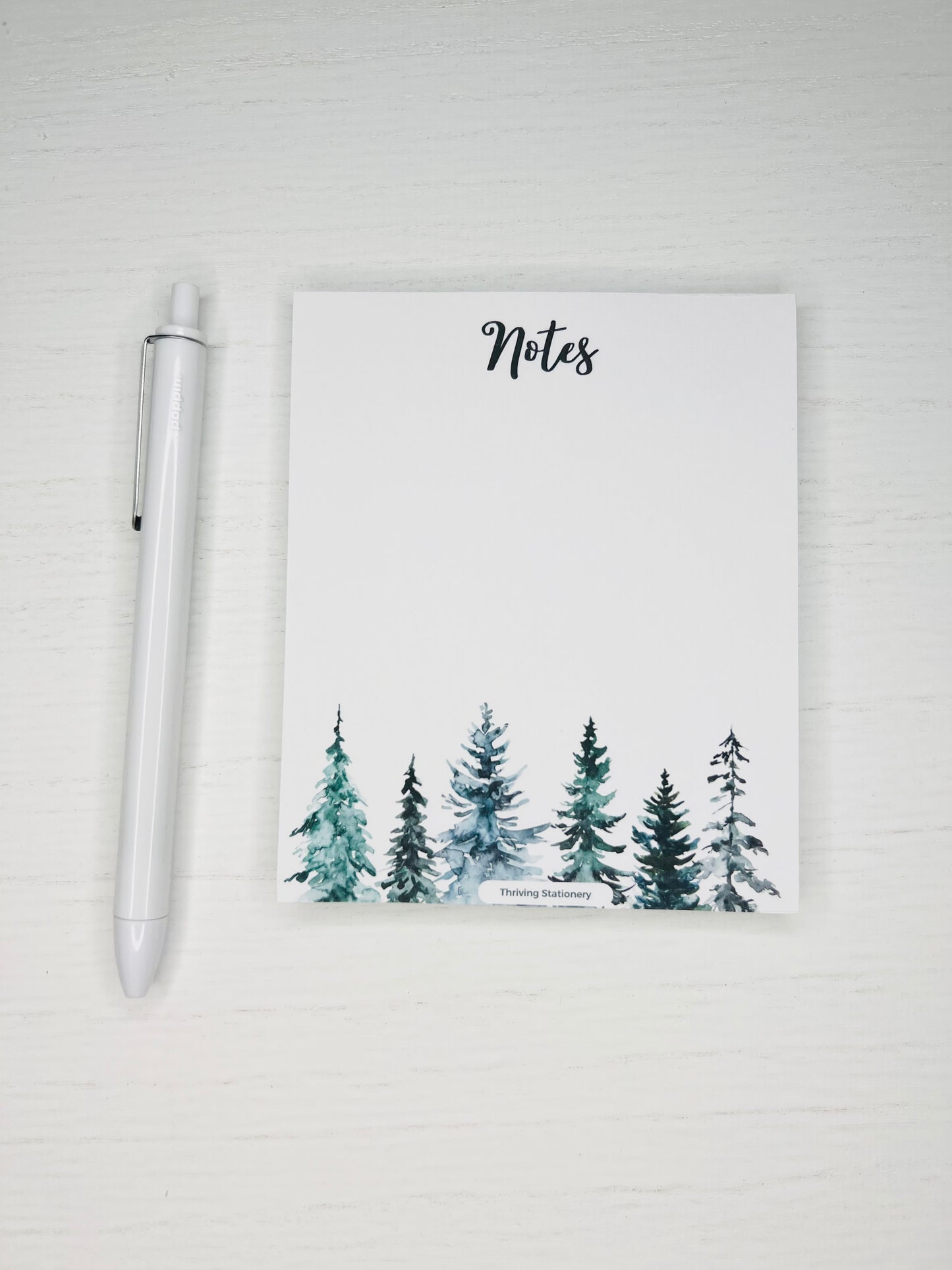 A blank notepad with a winter tree design, accompanied by a white pen, placed on a white surface.