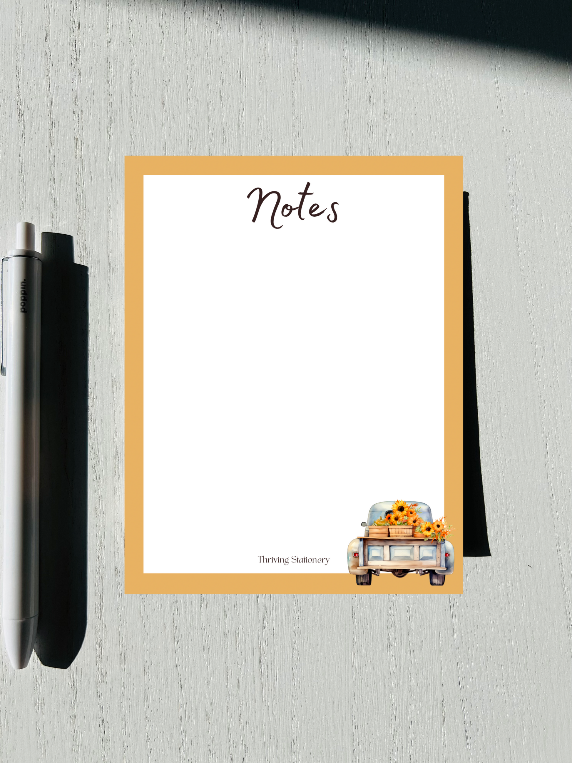 A notepad , featuring a design with autumn-themed graphics such as leaves and a pumpkin, alongside a pen and placed on a wooden surface.