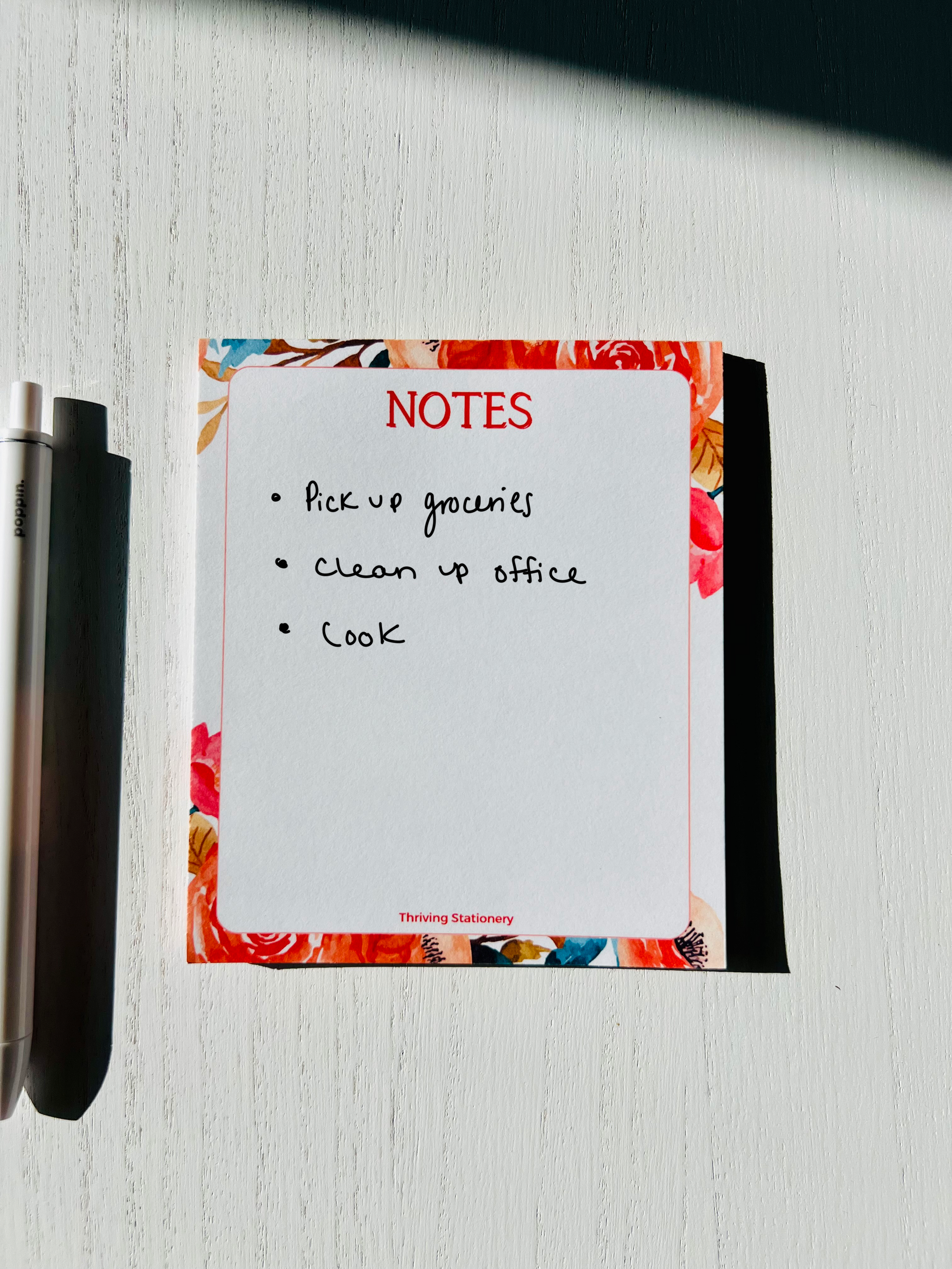 A mini notepad with a white, red, and natural color scheme, featuring a floral design on the cover, with a pen beside it.
