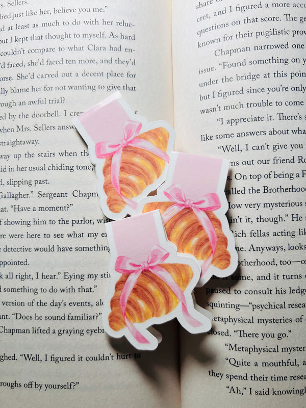 Two croissant-shaped bookmarks with pink ribbons on an open book.