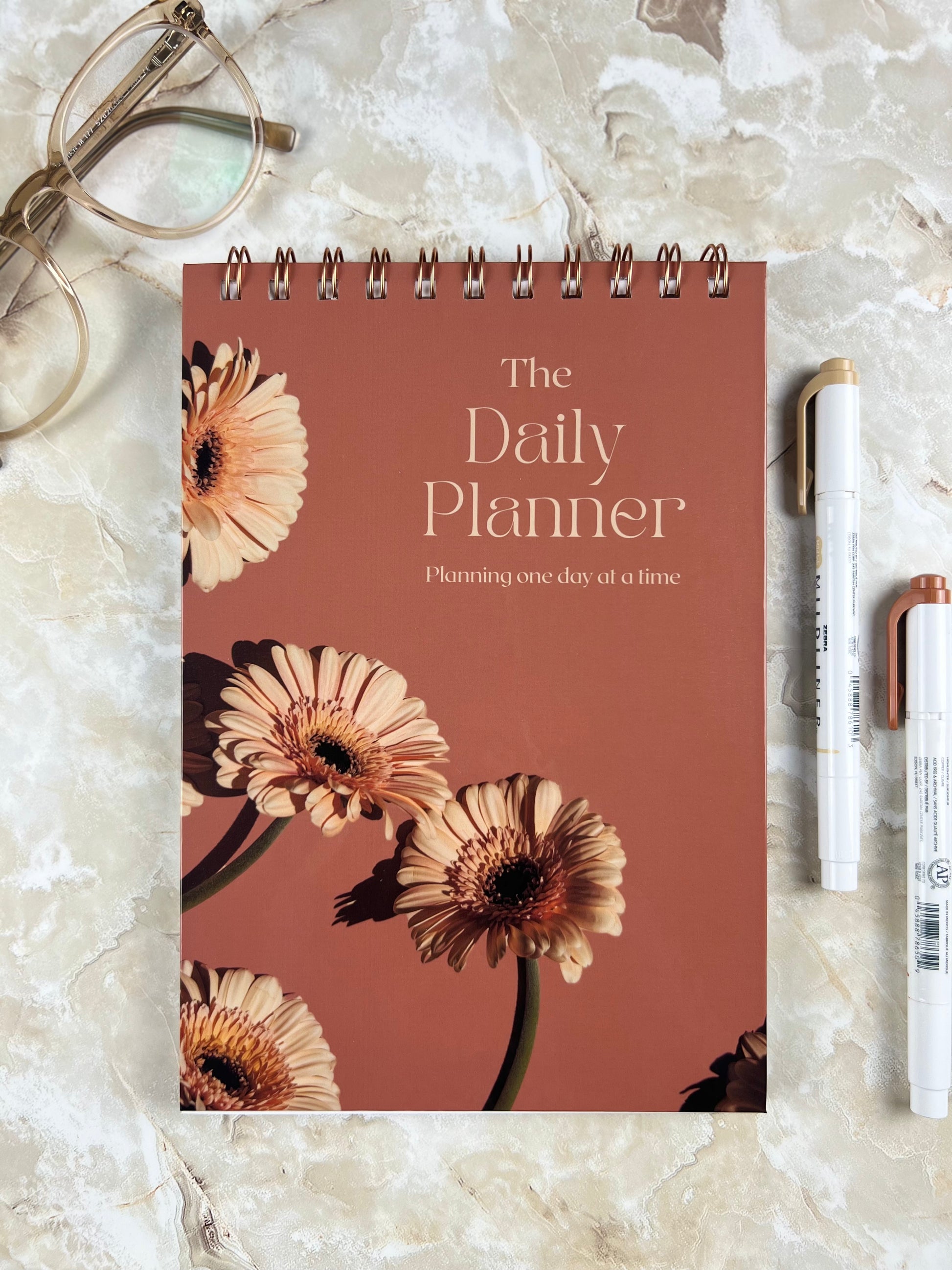 Daily planner with floral design on a marble surface with glasses and pens.