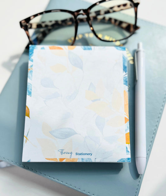 A blue-covered mini notepad with a floral pattern and the brand name 'Thriving Stationery' written on it, alongside a white pen and glasses on a blue textured surface.