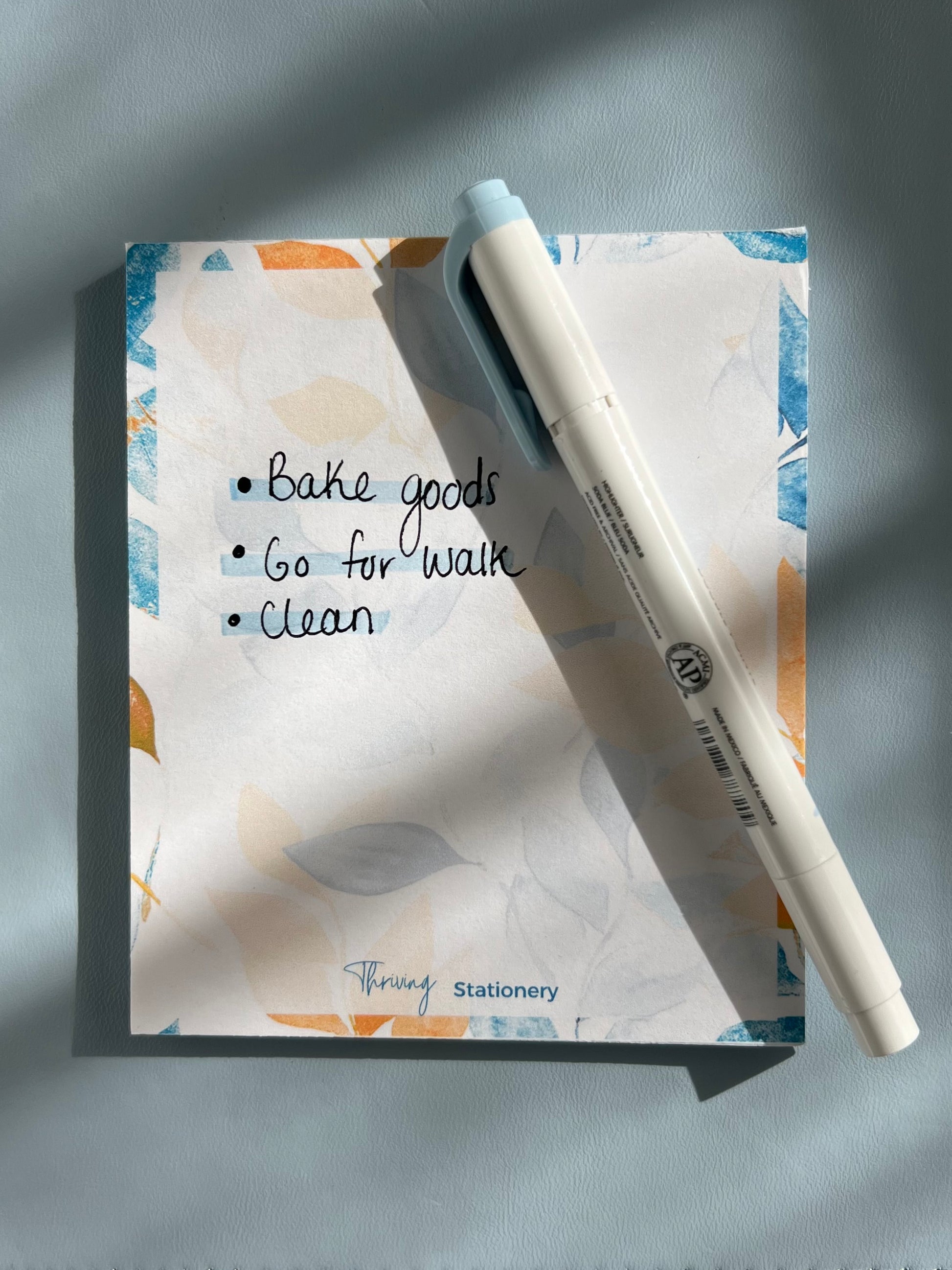 A blue-covered mini notepad with a floral pattern and the brand name 'Thriving Stationery' written on it, alongside a white pen and glasses on a blue textured surface.