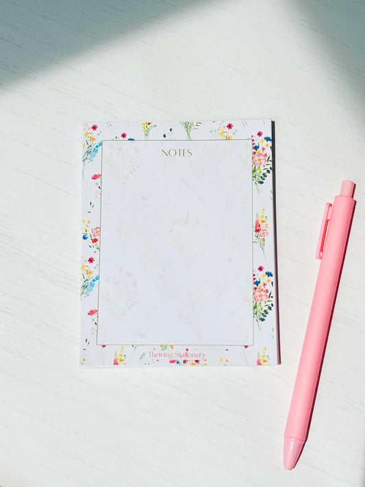 A floral-patterned blank notepad with a pink pen, labeled 'NOTES' on the cover, placed on a white surface.