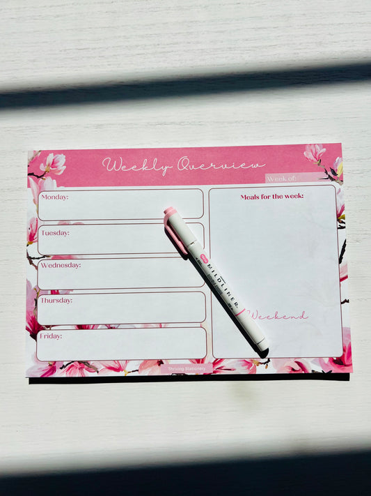 A pink weekly planner desk pad with floral borders and cherry blossom designs, featuring a white pen and marked with 'Weekly Review' in a flower-like font on the cover.
