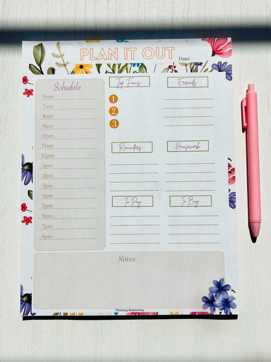 A white daily planner notepad with a floral design on the pages, accompanied by a pink pen.