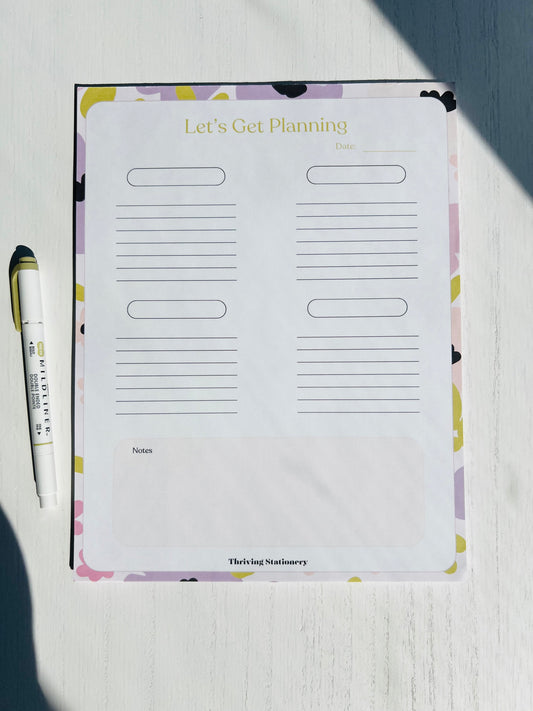 A large daily planner notepad with 'Let's Get Planning' titled on the cover, featuring a patterned design and a pen lying next to it.