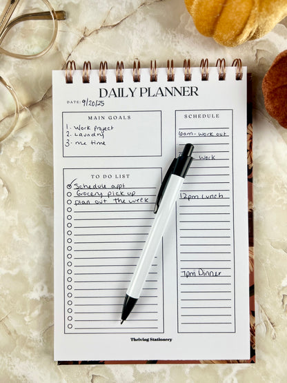 Fall Daily Planner with autumn floral cover and spiral binding for daily goals and scheduling with a pen for scale