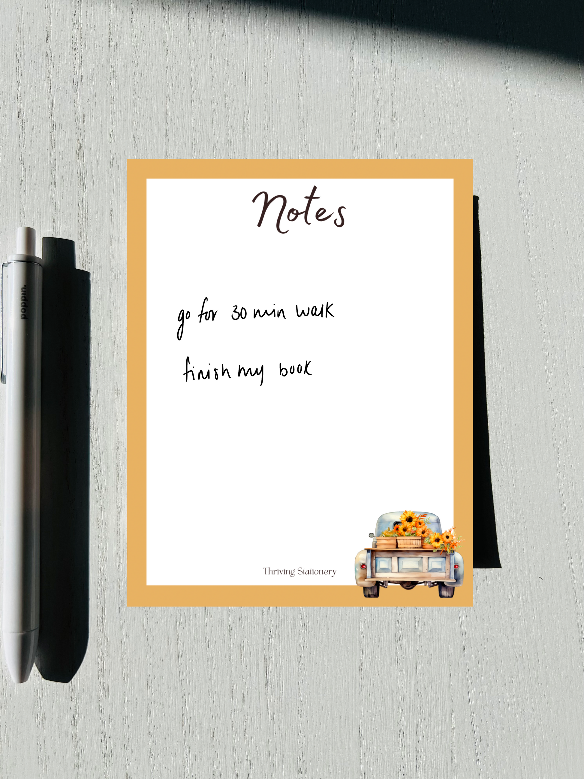 A notepad written, featuring a design with autumn-themed graphics such as leaves and a pumpkin, alongside a pen and placed on a wooden surface.