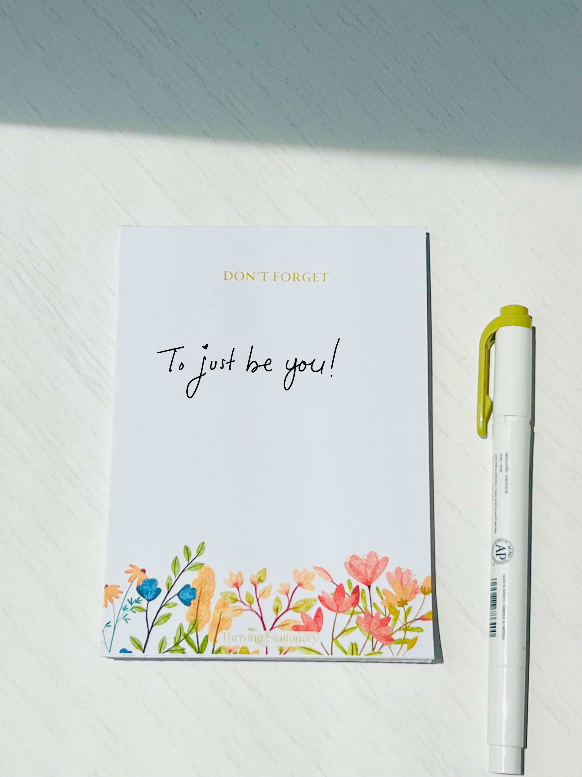 Minimal white notepad with soft watercolor floral illustrations along the bottom and “DON’T FORGET” in gold text at the top. Branding reads “Thriving Stationery.