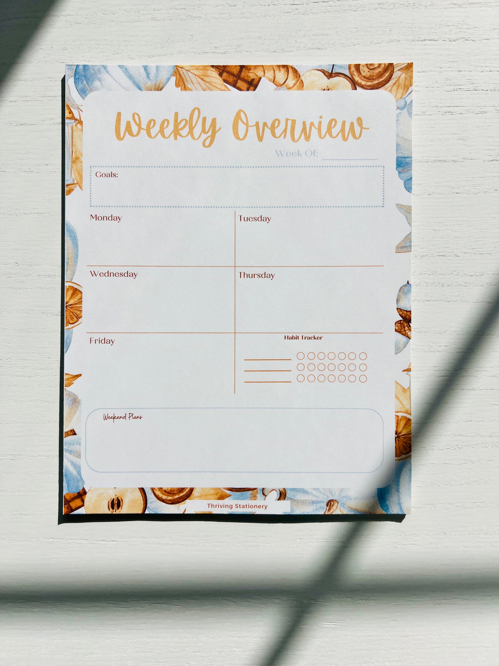 A weekly planner notepad with a fall pumpkin floral border and a title 'Weekly Overview' at the top, along with sections for Monday to Saturday and a blank Sunday space, placed on a surface with a light shadow falling over it.