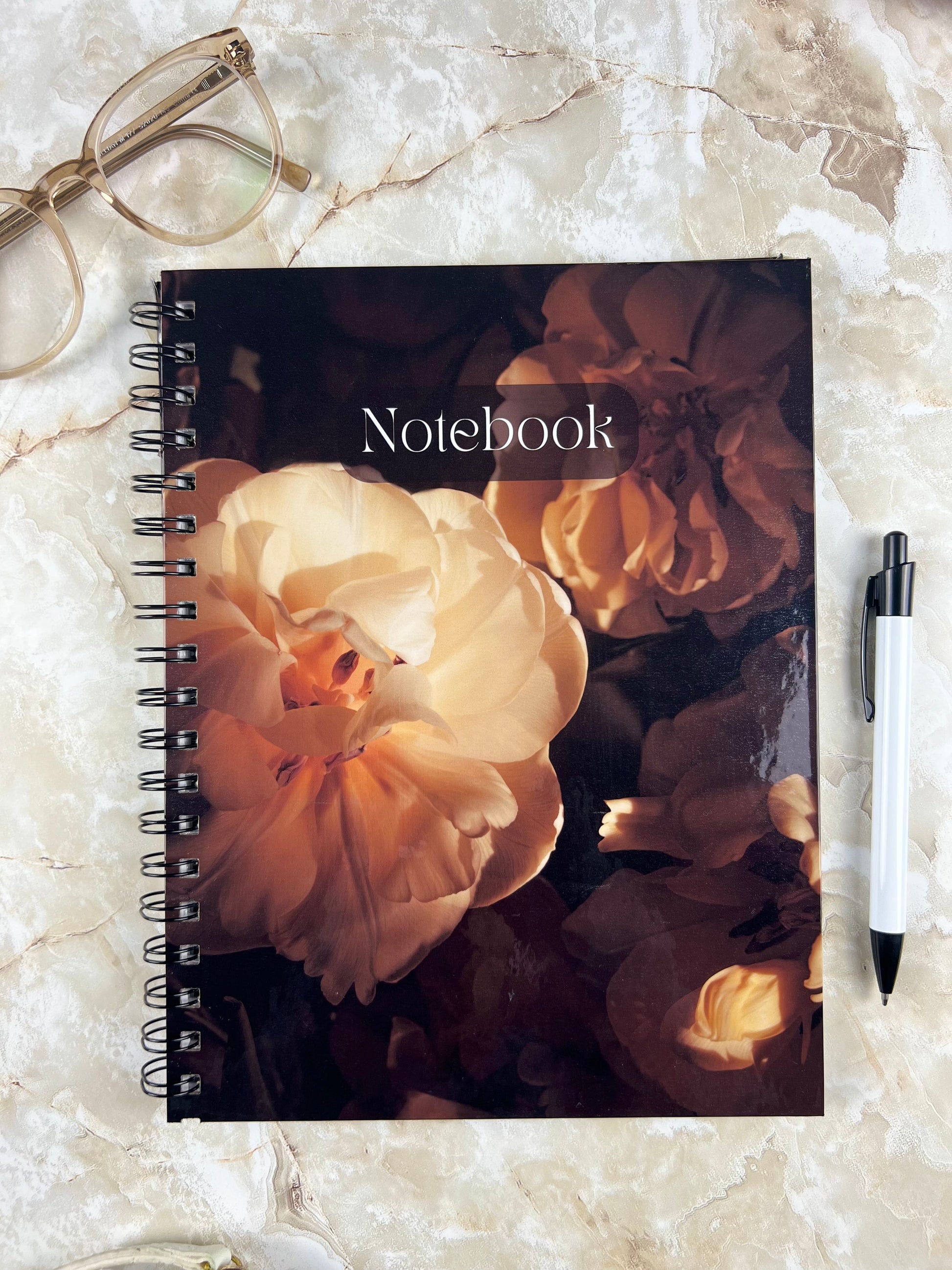 Floral notebook on a marble surface with glasses and a pen.