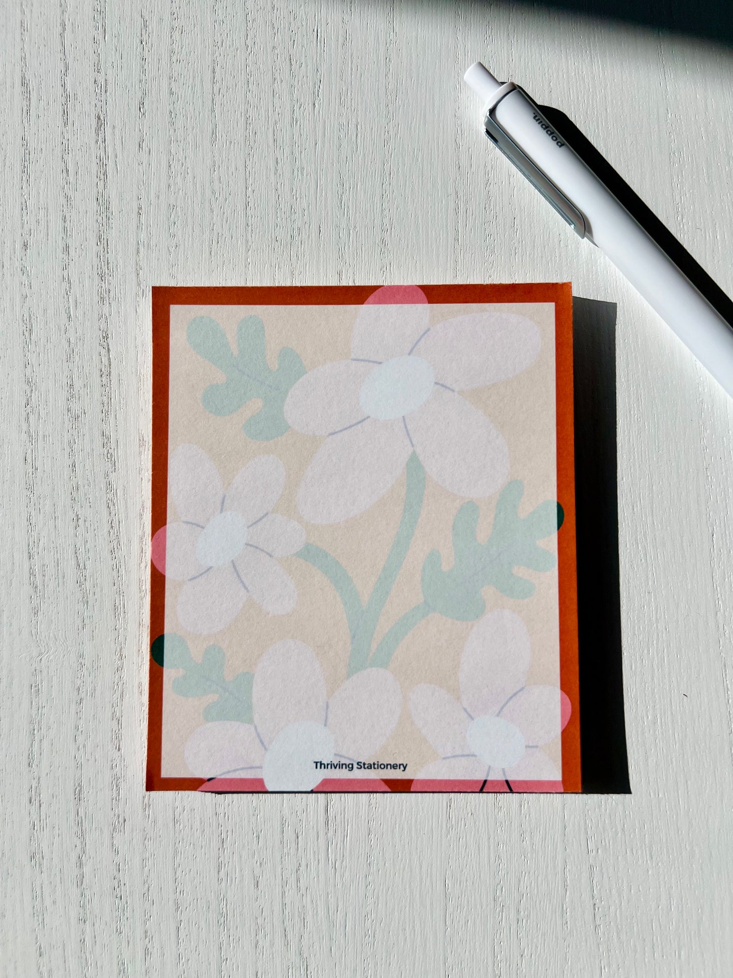 A mini notepad with a white, pink, and natural color scheme, featuring a floral pattern and the brand name 'Thriving' at the bottom.