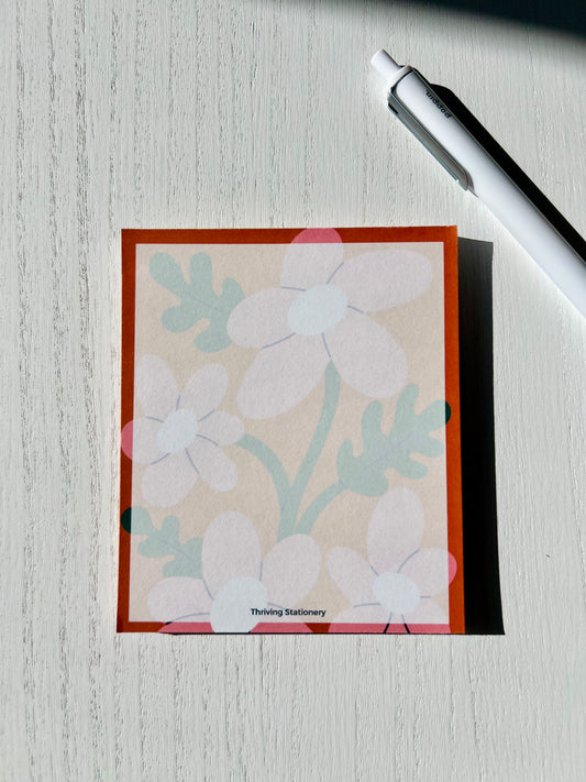 A mini notepad with a white, pink, and natural color scheme, featuring a floral pattern and the brand name 'Thriving' at the bottom.