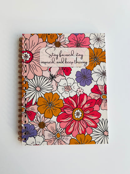 A spiral journal with a floral design cover in pink, orange, and purple petals, with a quote on the cover.