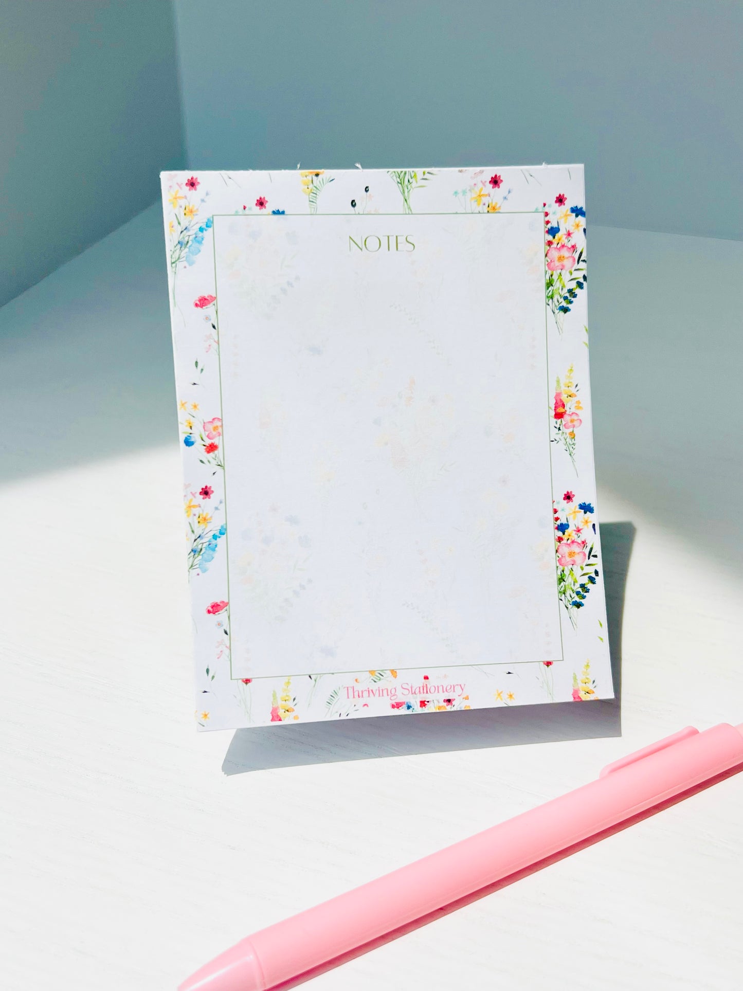 White notepad bordered with vibrant watercolor wildflowers. The word “NOTES” is printed at the top in a soft green serif font. Branding at the bottom reads “Thriving Stationery.”