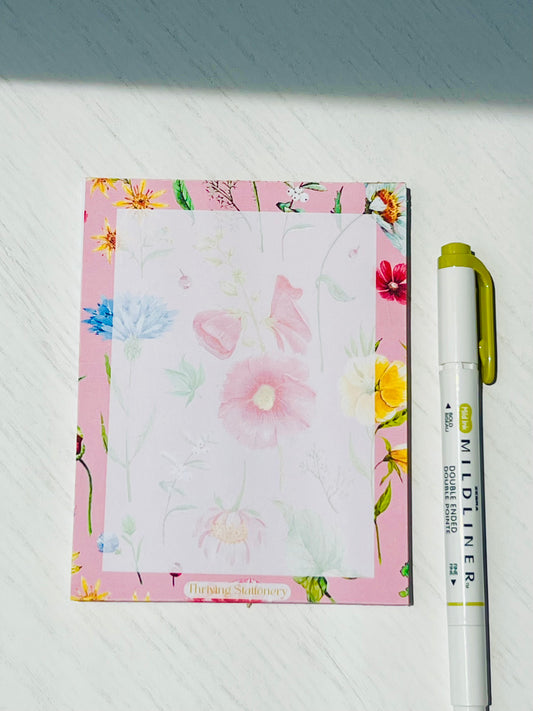 Floral notepad with soft watercolor wildflowers on a blush pink background. “Thriving Stationery” logo at the bottom center.