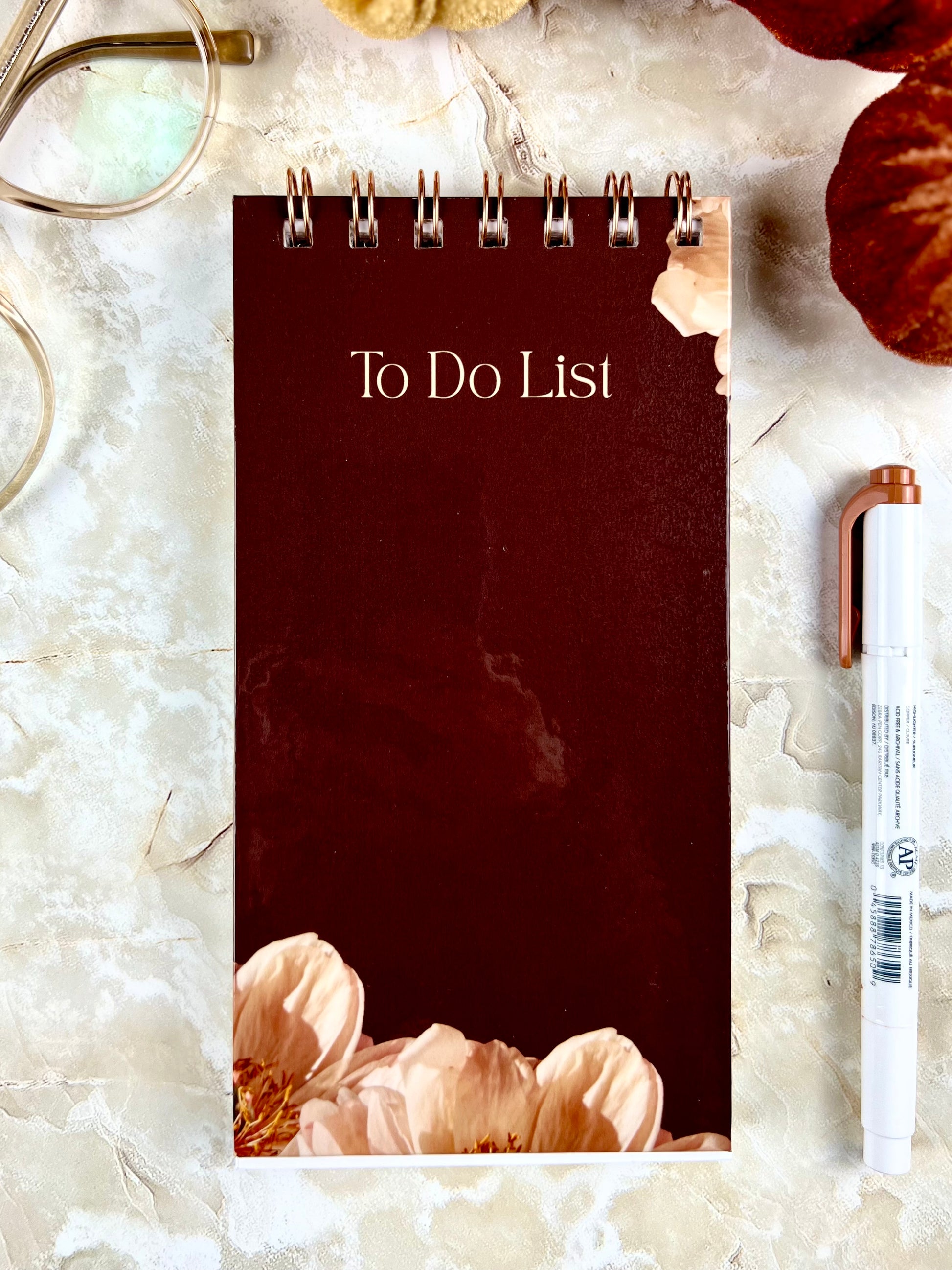 Maroon 'To Do List' notebook with floral design on a light marble surface with a pen and glasses.