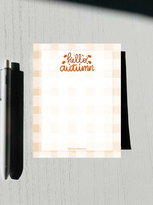 A Hello Autumn lined notepad with a checkered pattern, placed on a desk next to a pen.