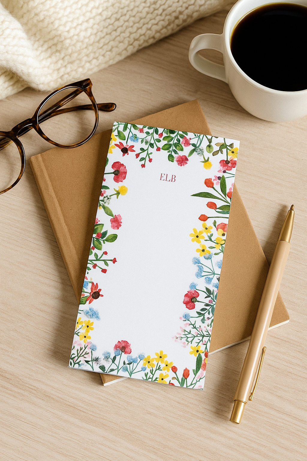 Floral notebook with 'ELB' initials on a wooden surface with a cup of coffee and glasses.