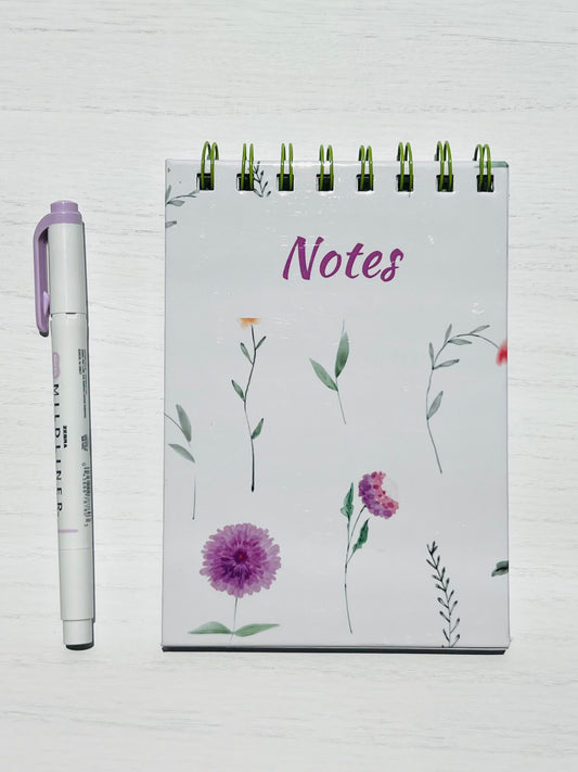 A small unlined notebook with a floral themed cover featuring a floral design, alongside a pen for scale.