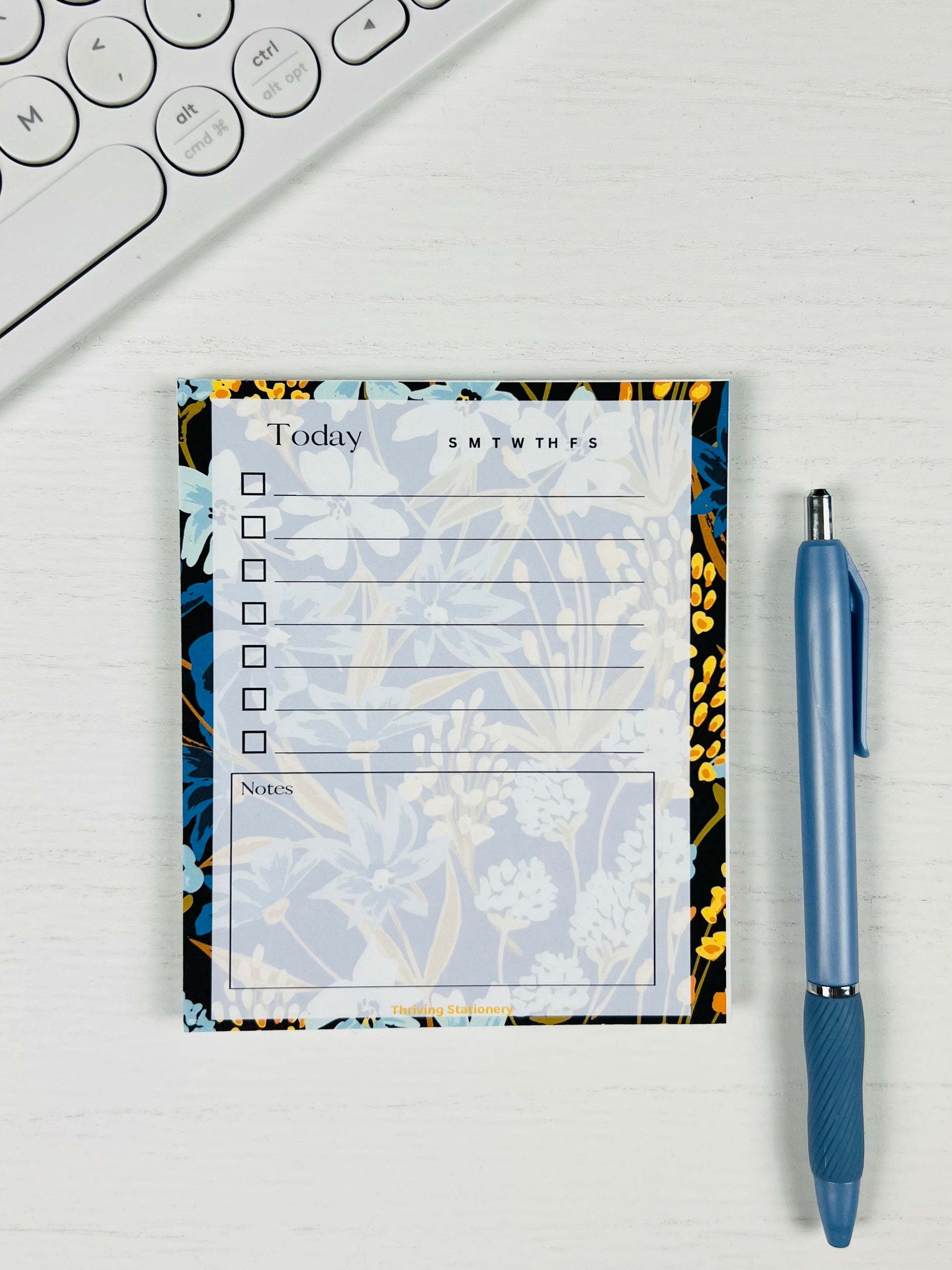 Navy Meadow Daily Planner Notepad with navy, blue, and gold floral border and daily checklist layout with a pen for scale