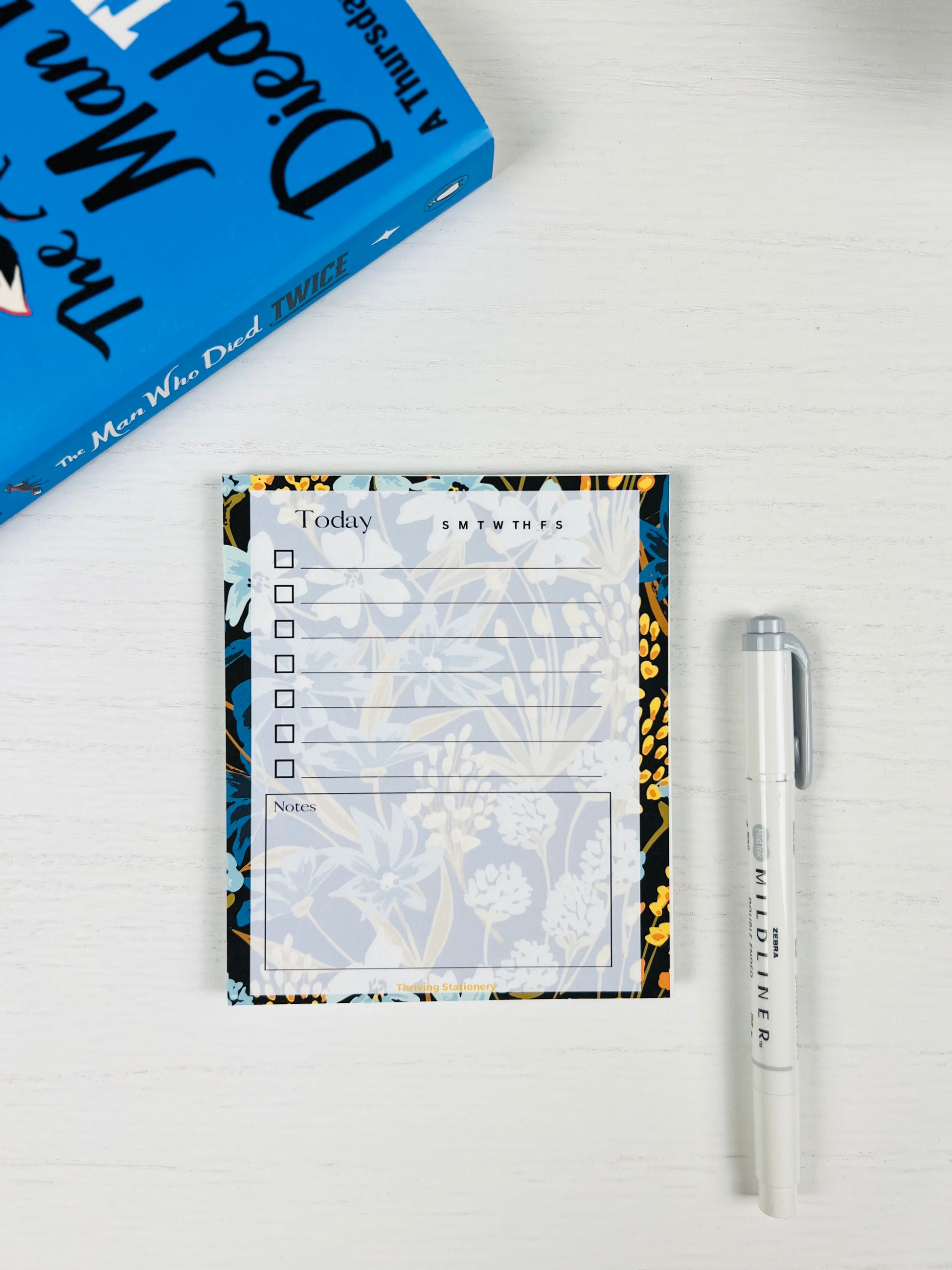 Navy Meadow Daily Planner Notepad with navy, blue, and gold floral border and daily checklist layout with a book and pen for scale