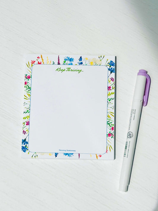 A multicolored blank notepad with a floral design on the cover, titled 'Keep Thriving', accompanied by a purple pen.