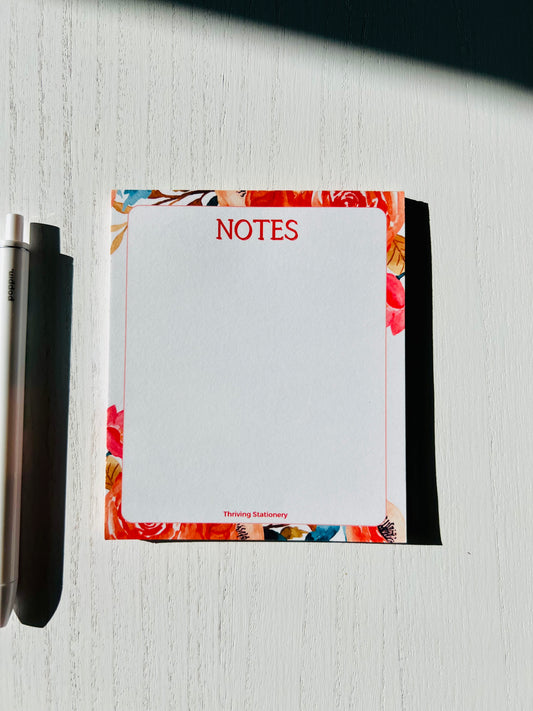 A mini notepad with a white, red, and natural color scheme, featuring a floral design on the cover, with a pen beside it.