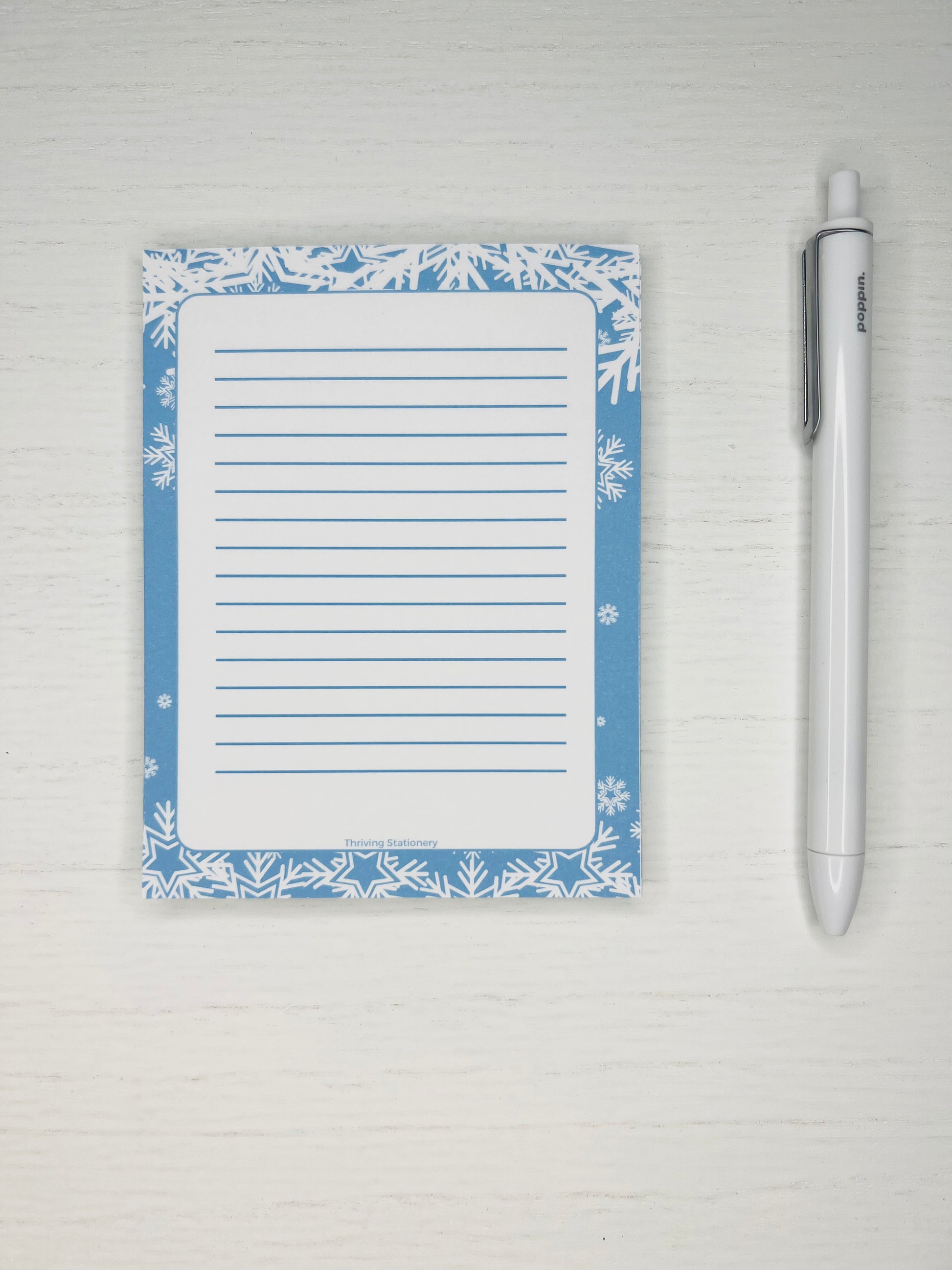 A blue and white lined notepad with a winter design, accompanied by a white pen, placed on a surface with a floral pattern.