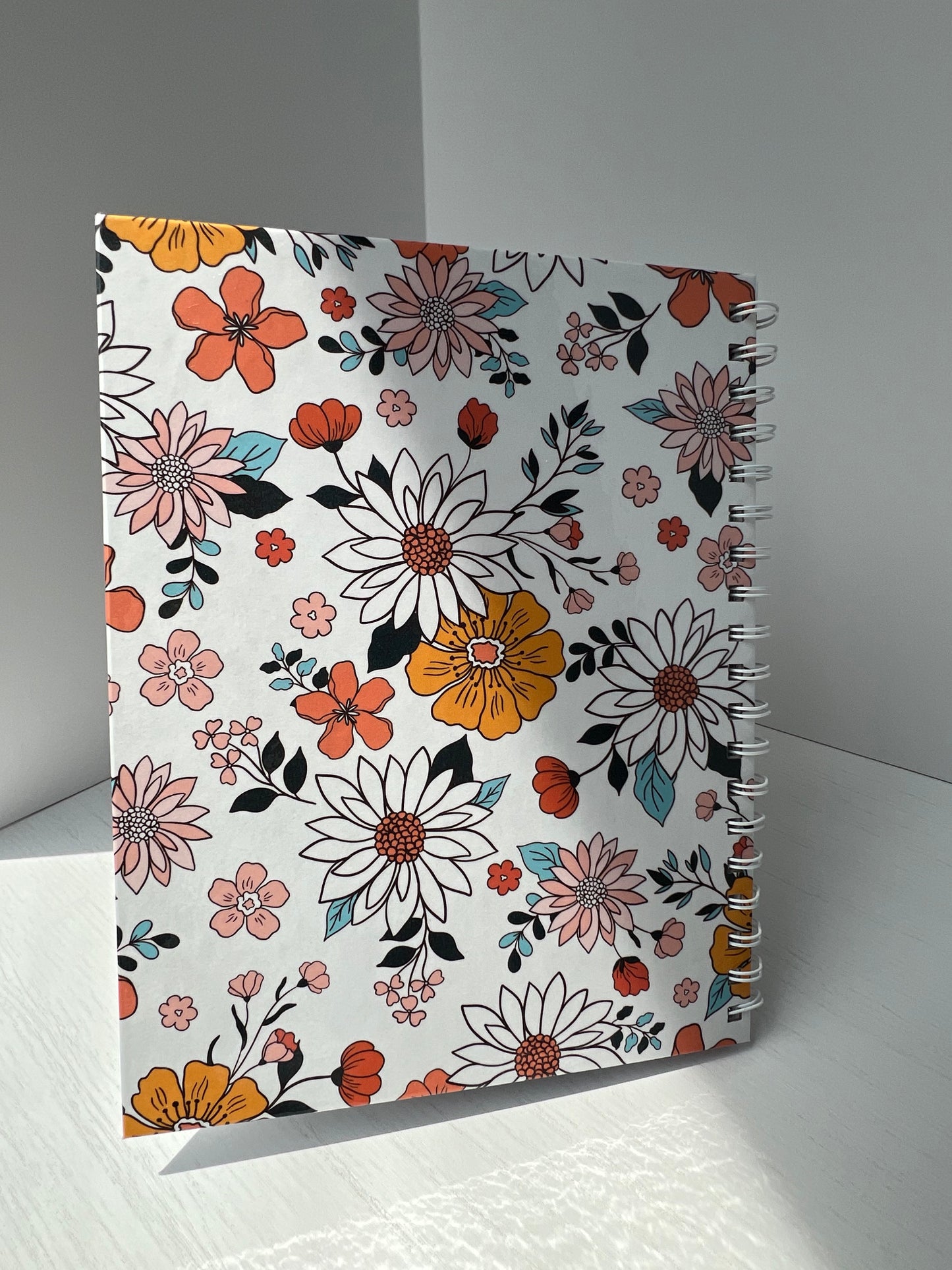 Floral spiral hardcover notebook with vibrant orange, blush, and white flowers on a white background. Labeled “Notebook” with “Thriving Stationery” branding.