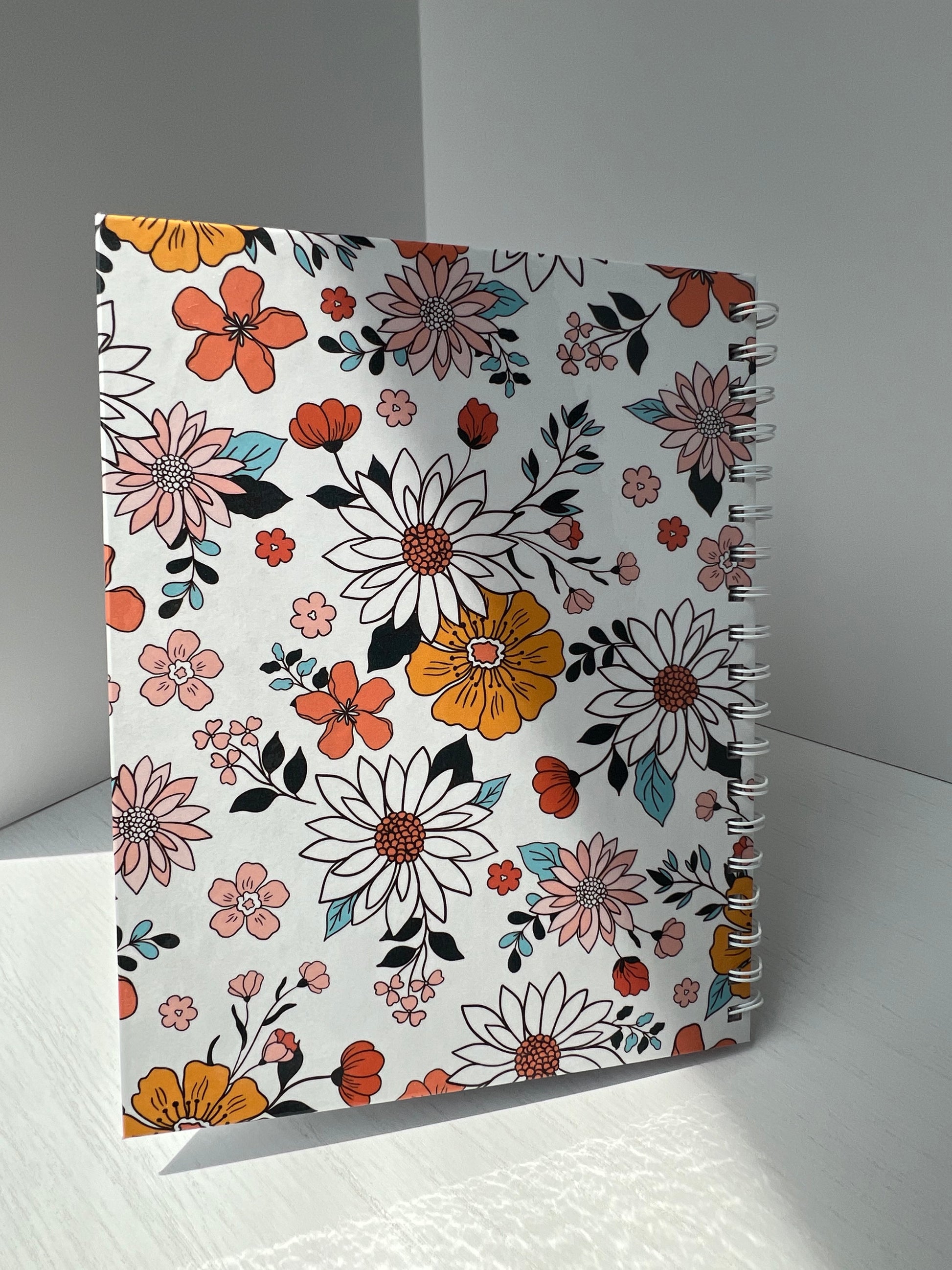 Floral spiral hardcover notebook with vibrant orange, blush, and white flowers on a white background. Labeled “Notebook” with “Thriving Stationery” branding.