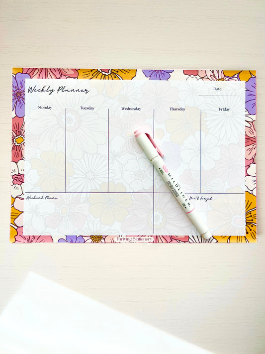A colorful floral-patterned weekly planner desk pad with a pen and a flower design on the cover page.